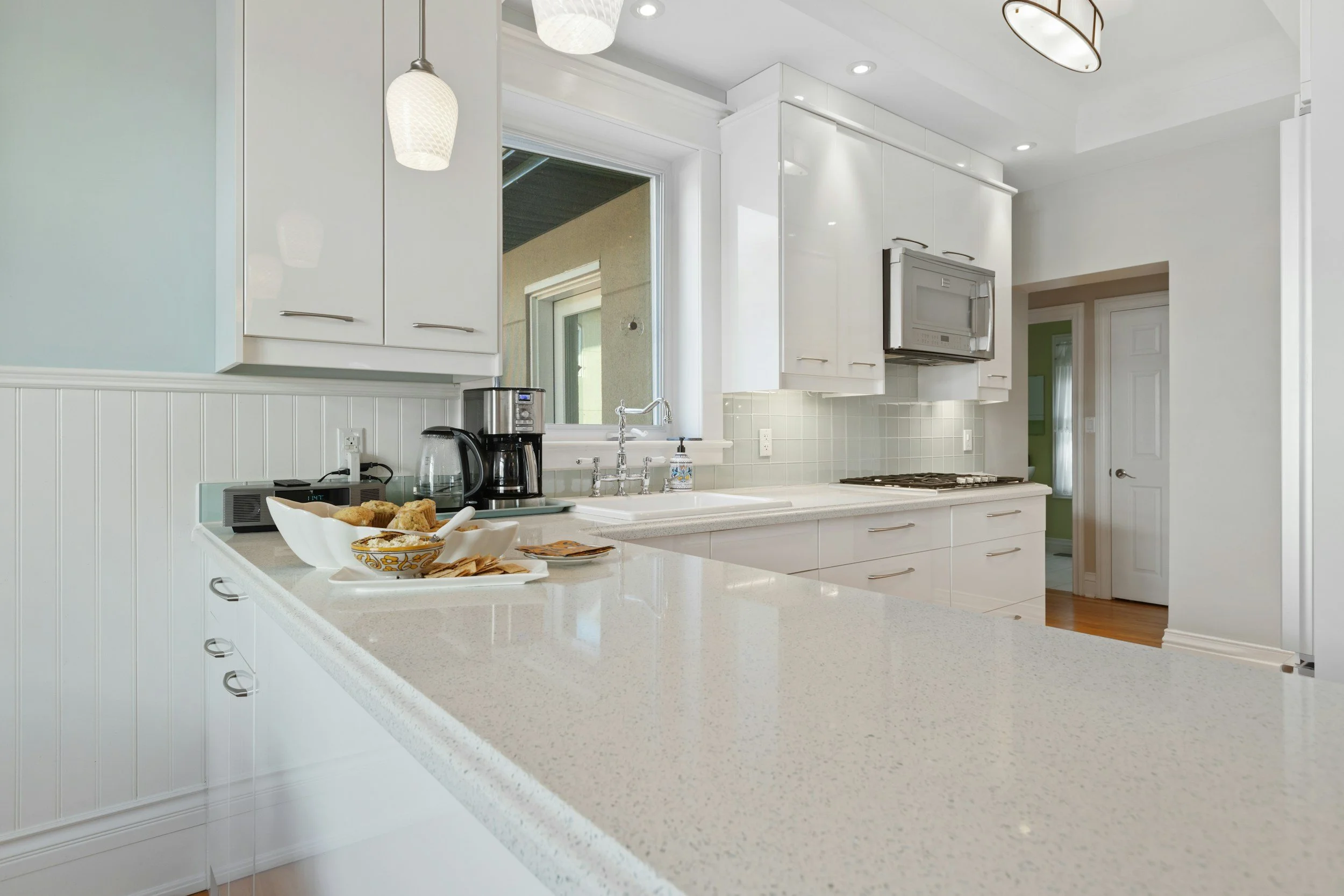 A modern kitchen with white cabinets, a speckled white countertop, a window above the sink, stainless steel appliances, and a breakfast bowl with muffins on the counter.