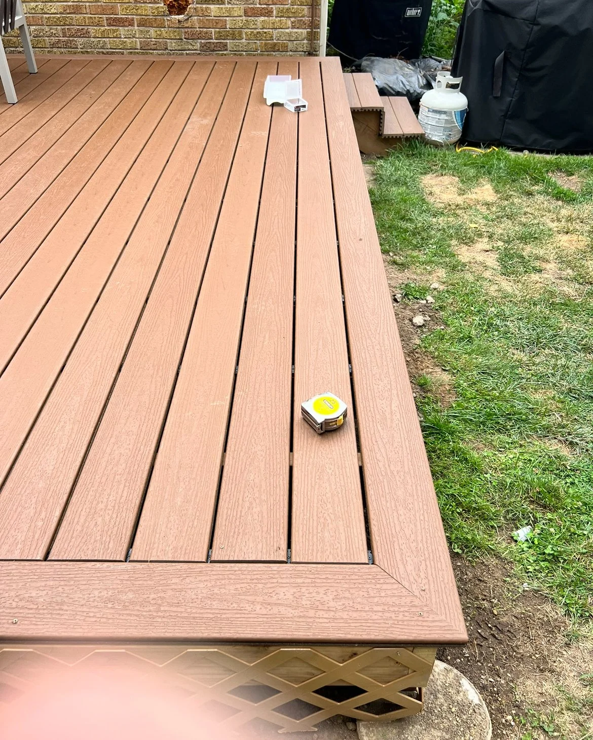 Newly installed wooden deck with a tape measure on it, adjacent to grass and a brick wall, with a propane tank and outdoor equipment nearby.