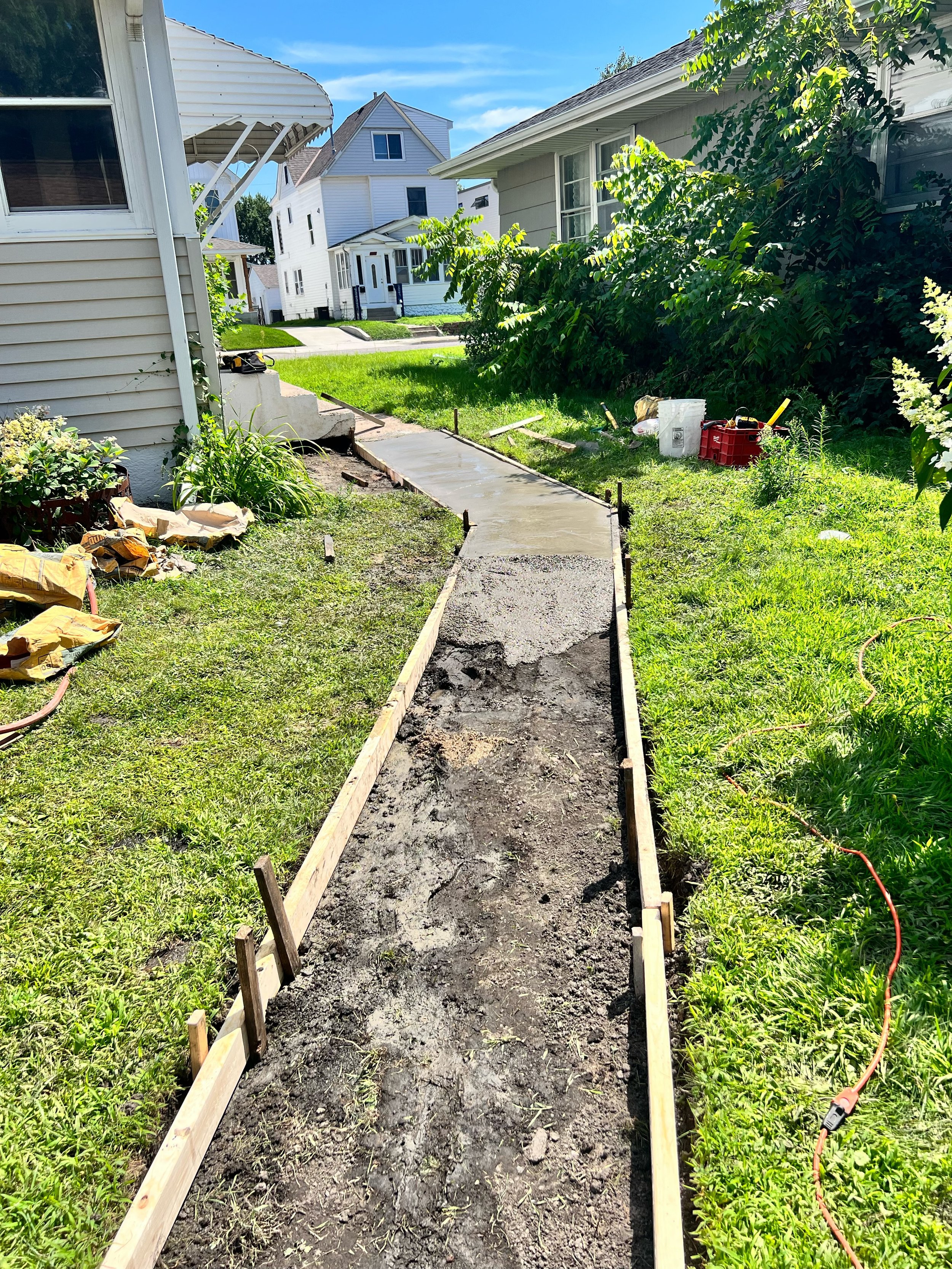 A newly poured concrete sidewalk in a backyard, with wooden forms on the sides, surrounded by grass, plants, and house exteriors on a sunny day.