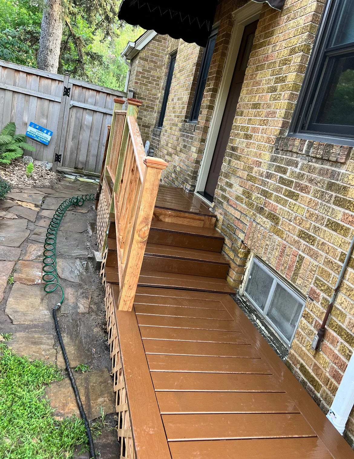 Backyard patio area with newly installed wooden stairs and deck, brick house exterior, garden hose on stone pathway, and a wooden fence with a political campaign sign.