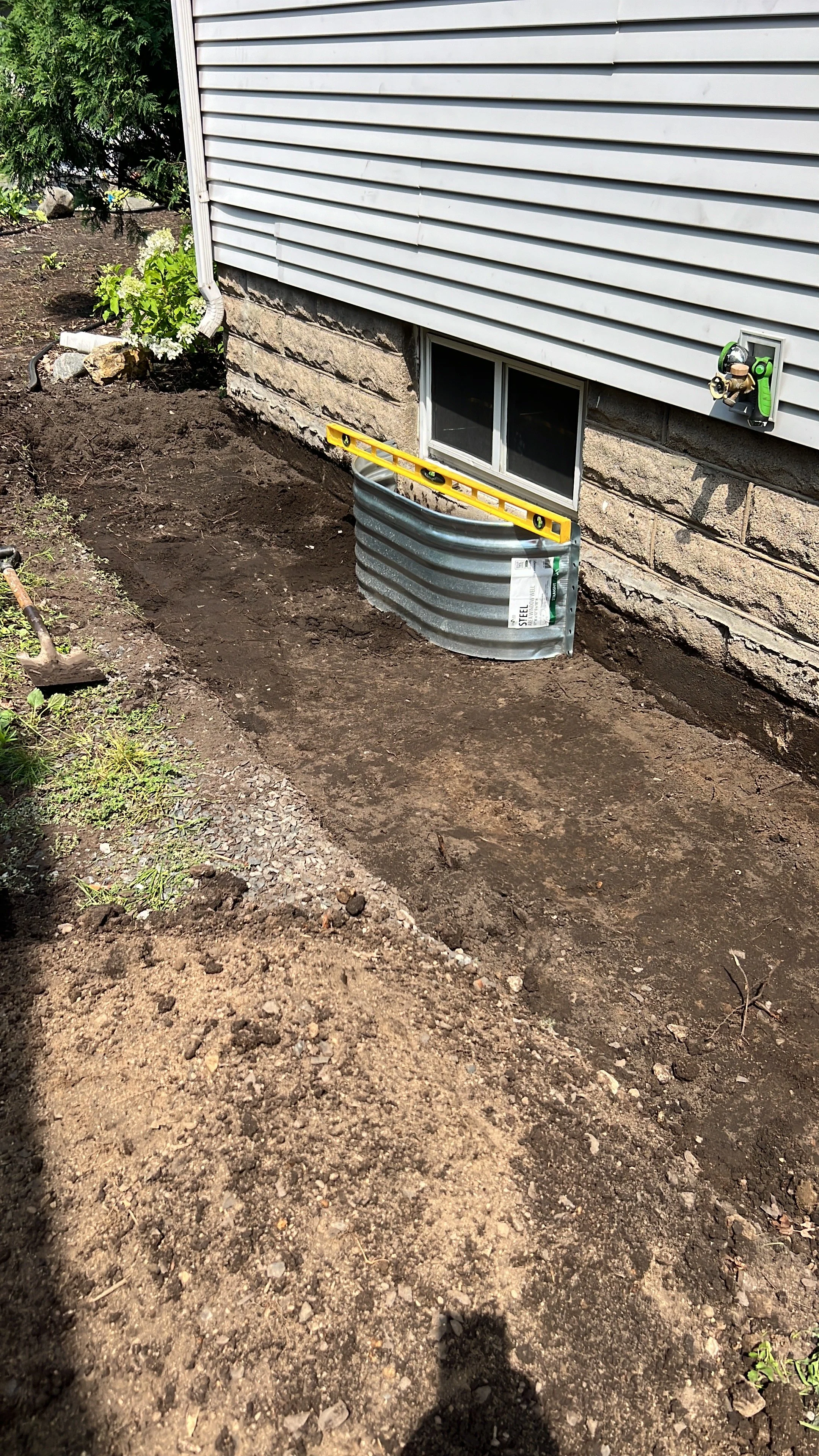 Excavated area of soil beside a house foundation with a small window, a ledge, a garden hose, and some plants, indicating ongoing construction or landscaping work.