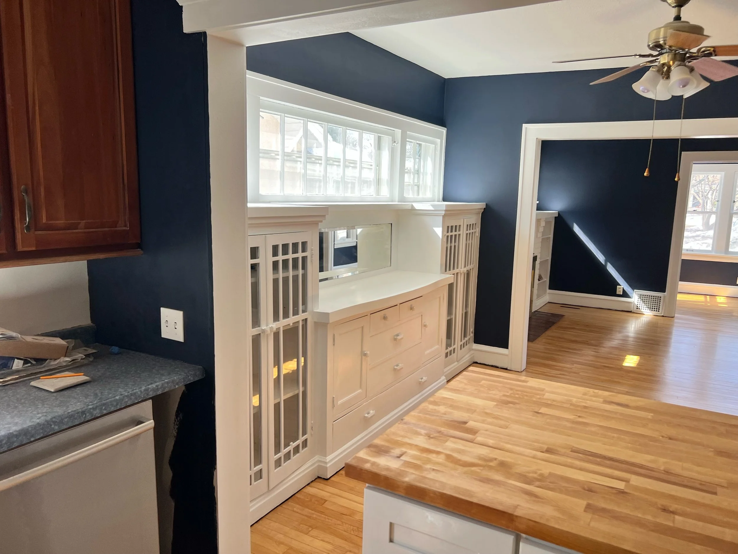 View of a living room with blue walls, a large window, and white built-in cabinets. A ceiling fan with lights is visible, and there is hardwood flooring.
