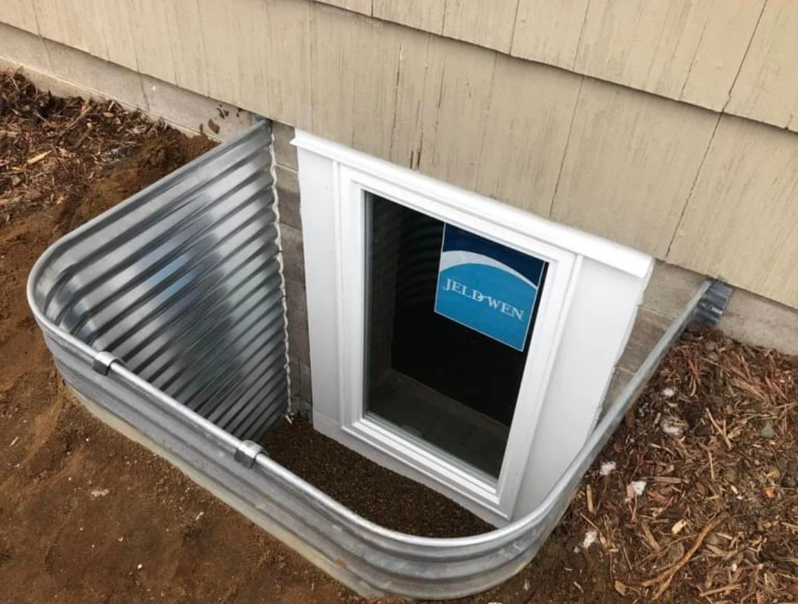 A basement window with a white frame and a sticker that says 'JELD WEN', surrounded by exterior wall siding, with a metal storm window and a small partially filled window well.