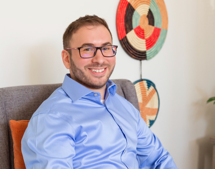 Smiling man wearing glasses and a blue shirt, sitting on a gray couch with an orange pillow, in a room with colorful wall art.