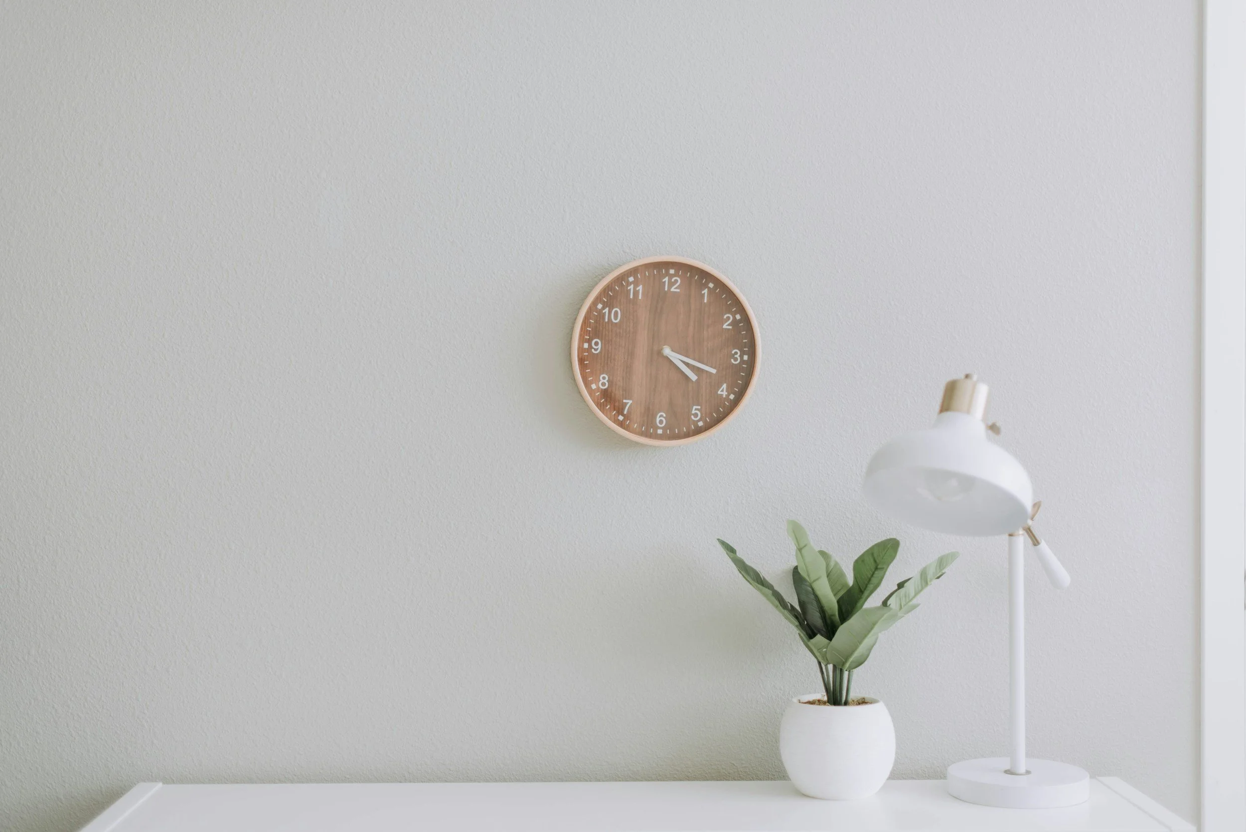 A wall clock with a wooden frame showing the time as 4:20 above a white vases with a green plant, a white desk lamp, on a white table against a light-colored wall.