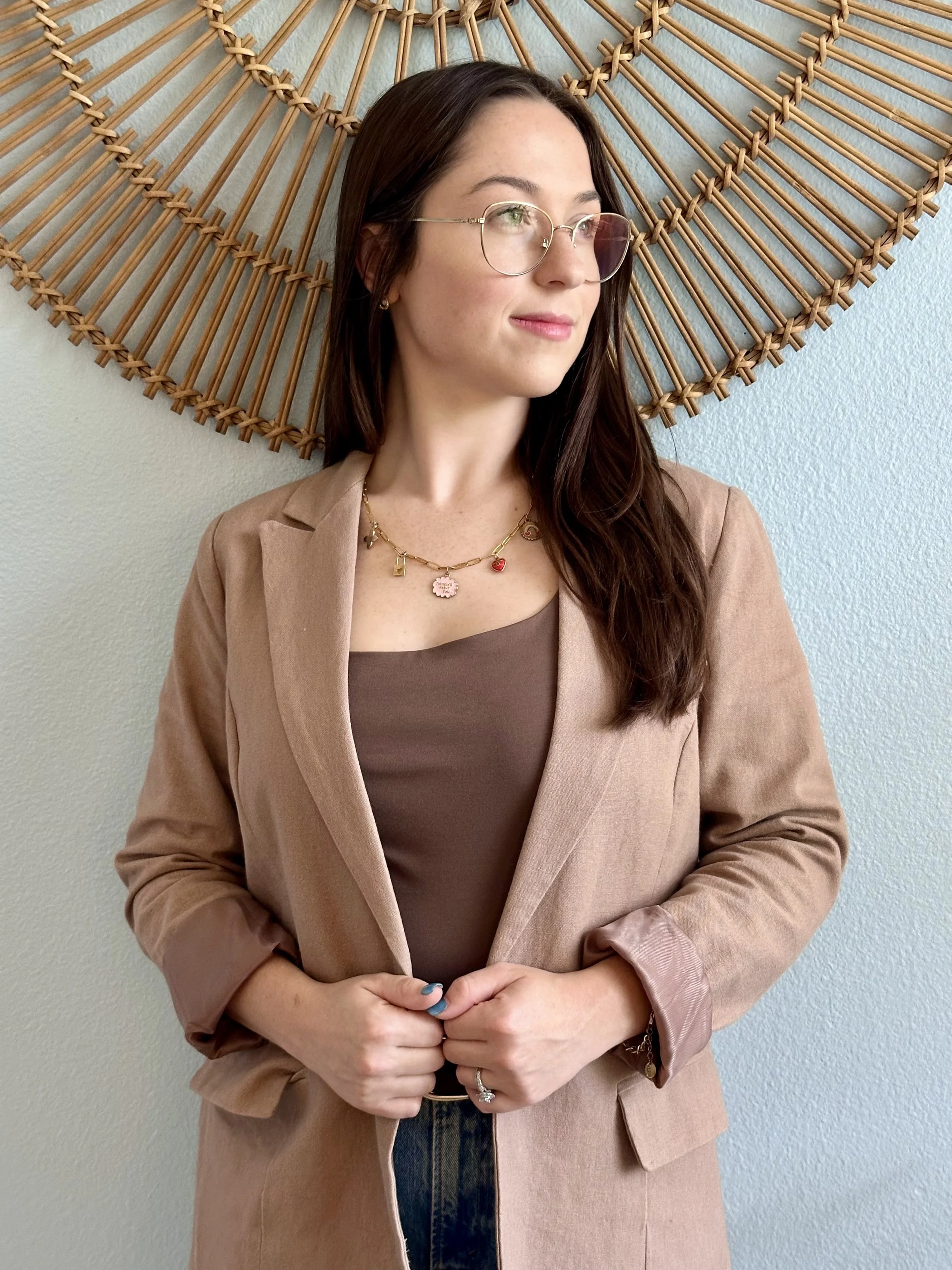 A woman with long brown hair and glasses, wearing a light brown blazer over a brown top, standing in front of a textured wall with a large decorative wooden star behind her.