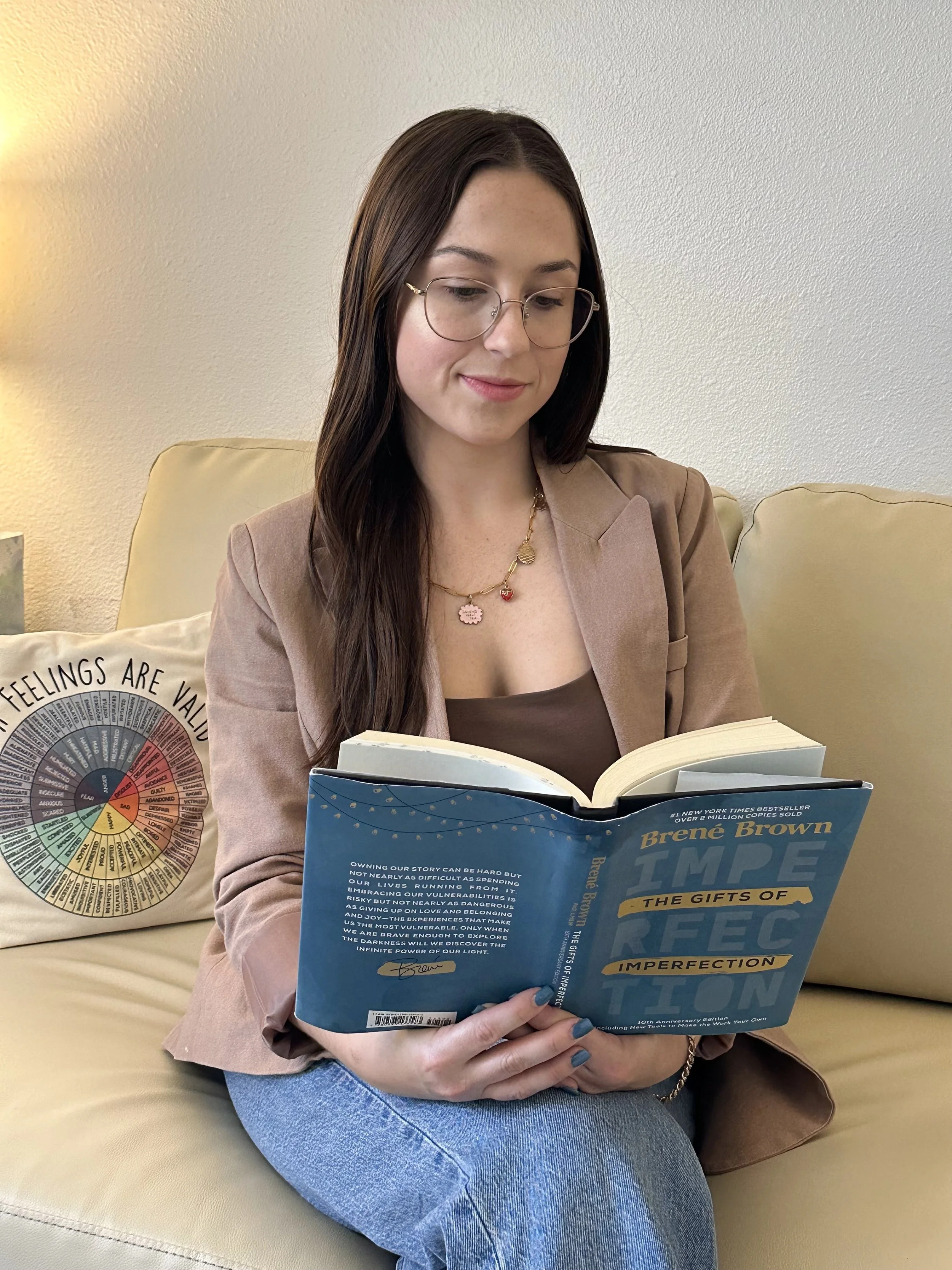 A woman sitting on a yellow couch reading a book titled "The Gifts of Imperfection" by Brené Brown. She has long dark hair, glasses, and is wearing a pink blazer and a necklace with a pink charm. There is a pillow behind her, part of which has a colorful wheel design and the text "Feelings are Valu" visible.