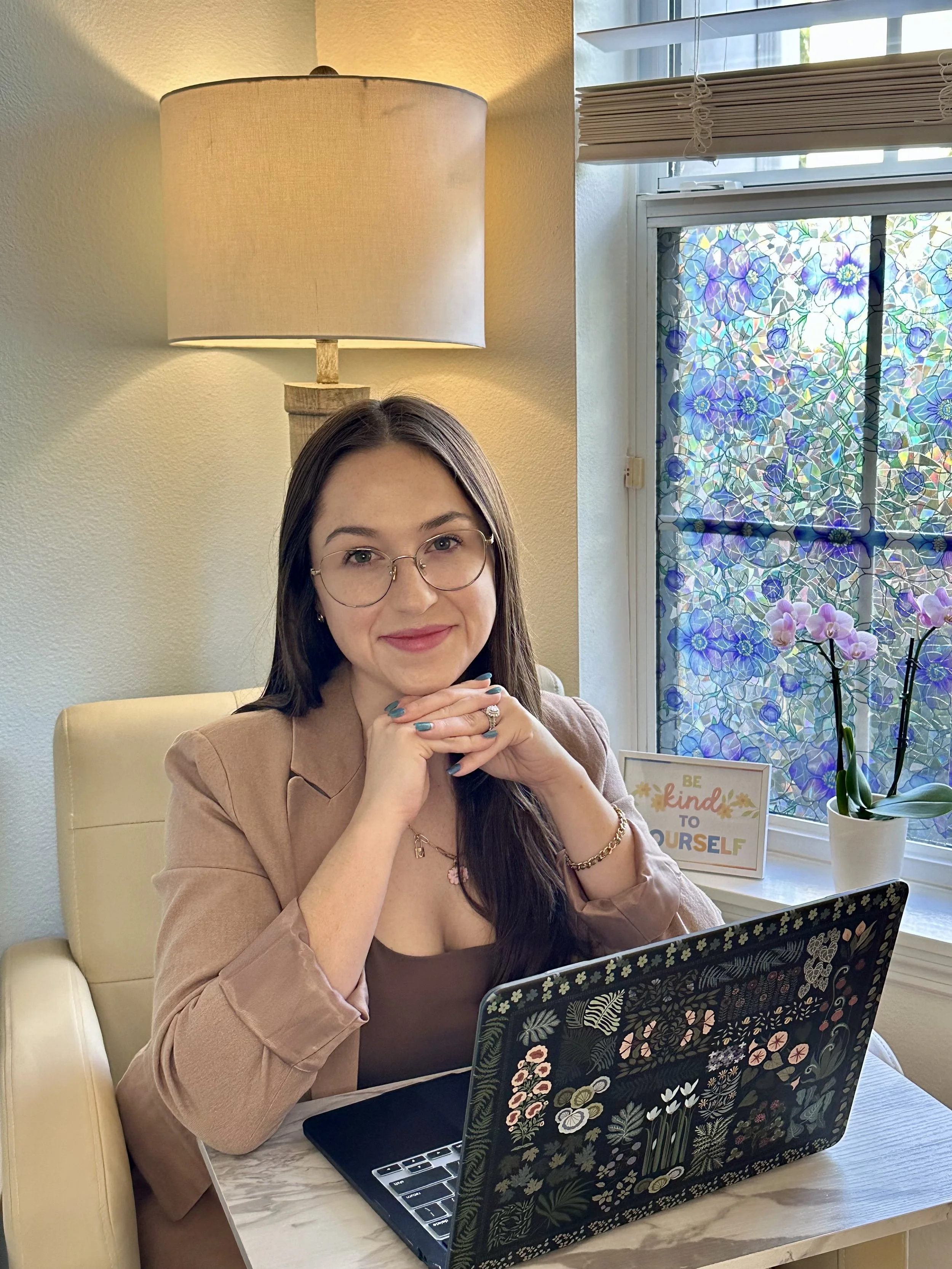 A woman sitting at a table working on a laptop with a floral design cover, in front of a window with stained glass and a sign that says 'Be kind to yourself.' She is wearing glasses, a beige blazer, and has long dark hair.