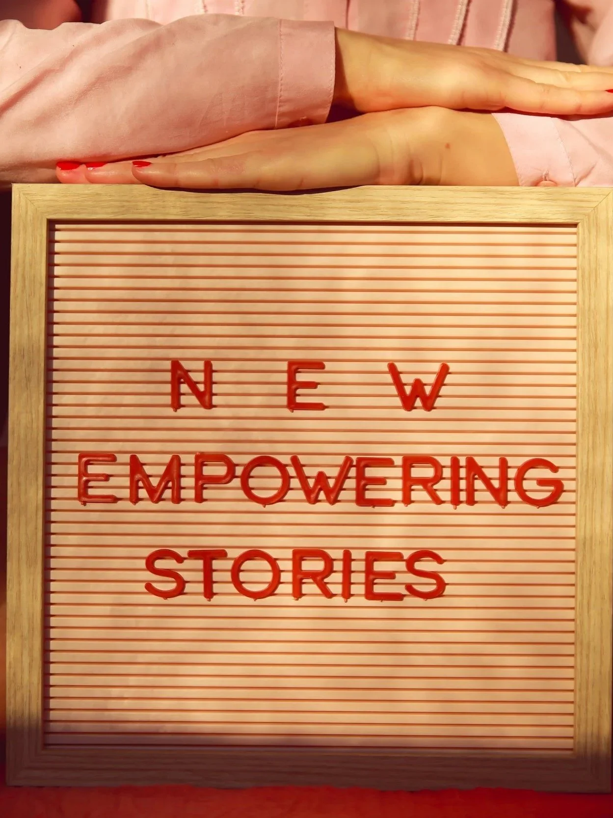 A person with pink nails and a pink long-sleeve top resting their hand on a wooden framed letter board. The letter board has red plastic letters spelling out "NEW EMPOWERING STORIES" on a pink striped background.