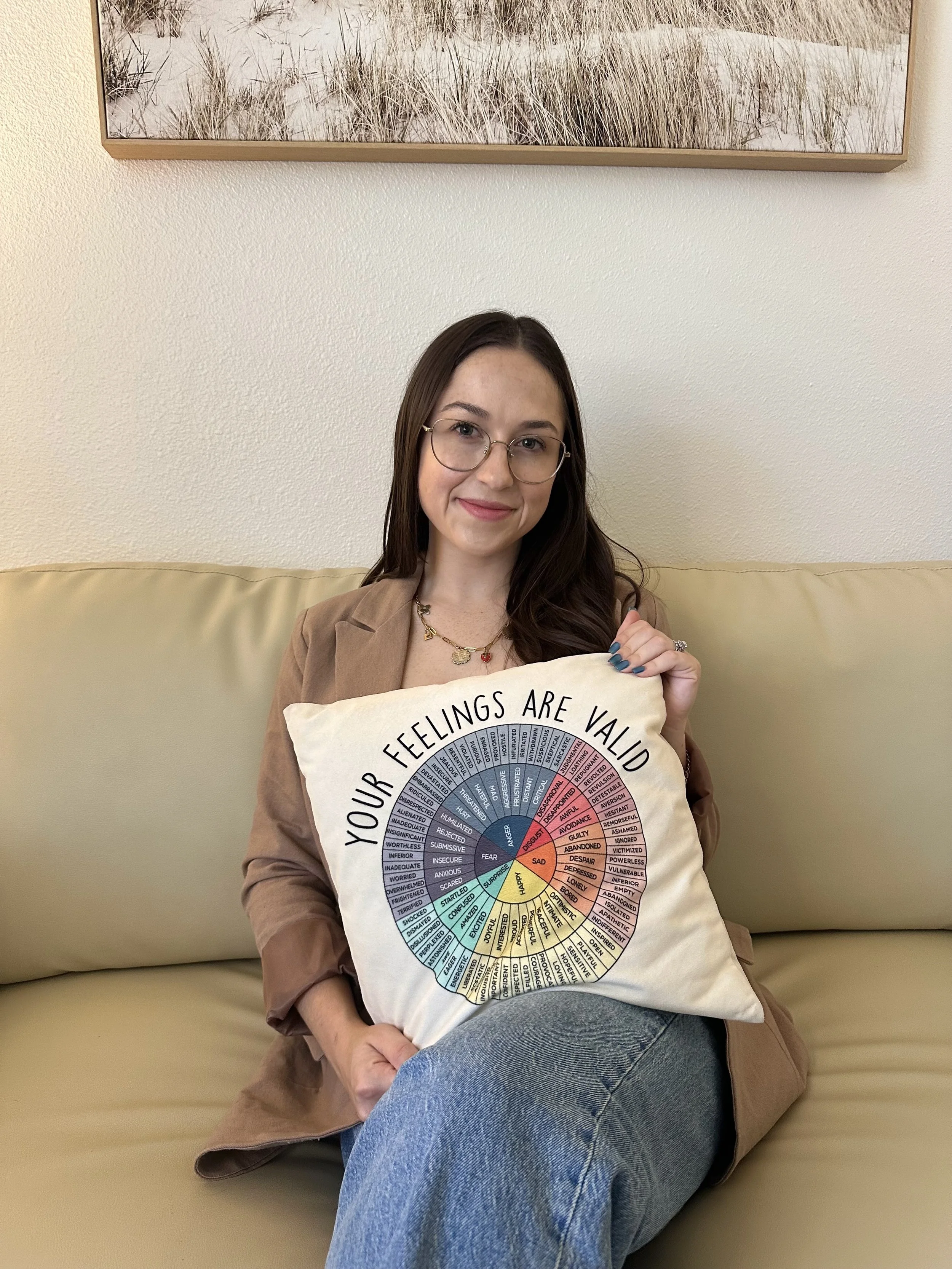 A young woman with glasses and long dark hair, sitting on a beige couch, smiling, and holding a pillow with a colorful emotional wheel chart and the words "Your feelings are valid" on it.