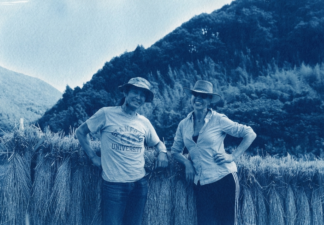 Two women standing in a wheat field with hills in the background, wearing hats and casual clothing, smiling.