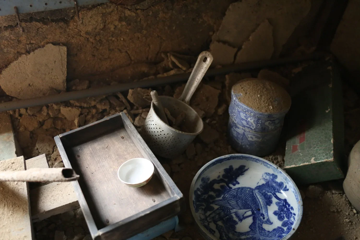 Old dusty kitchen space with a metal colander, a small white bowl, a decorated blue and white bowl, a metal container with dust, and a green box with a red and white label on the floor