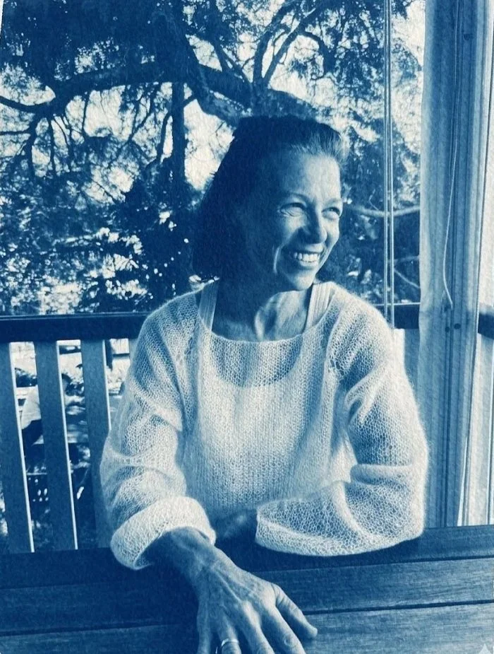 A woman sitting at a table outside, smiling and laughing, with large trees and a window in the background.