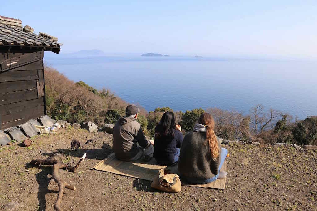 Three people sitting outdoors on a mat, overlooking a large body of water with distant islands, near a small wooden building and surrounded by trees and rocks.