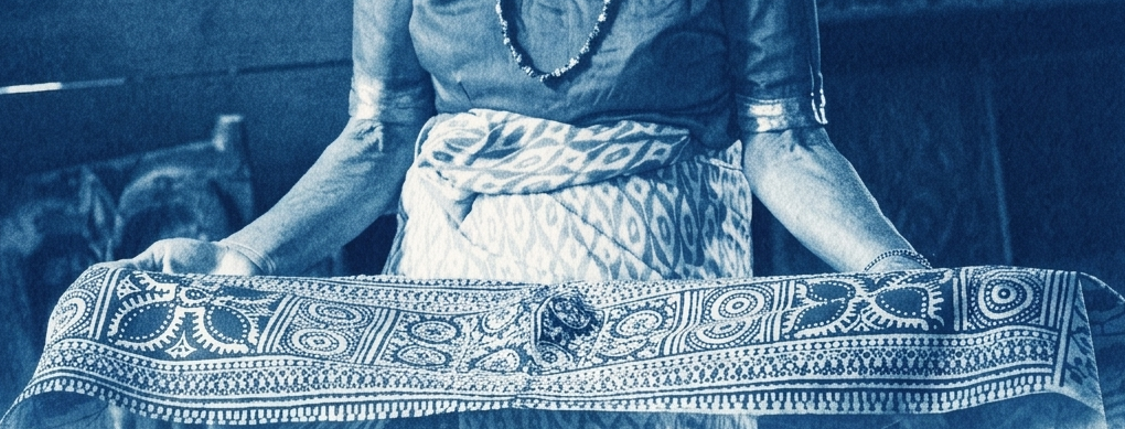 A person holding a patterned fabric or tablecloth, wearing a dress with a necklace, in a room with wooden furniture.