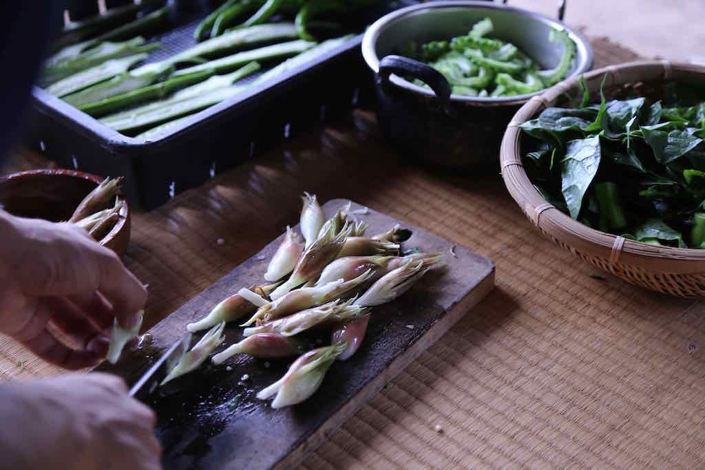 Hands preparing garlic cloves on a wooden cutting board surrounded by bowls of green vegetables, including Thai basil, and a tray of long green beans.