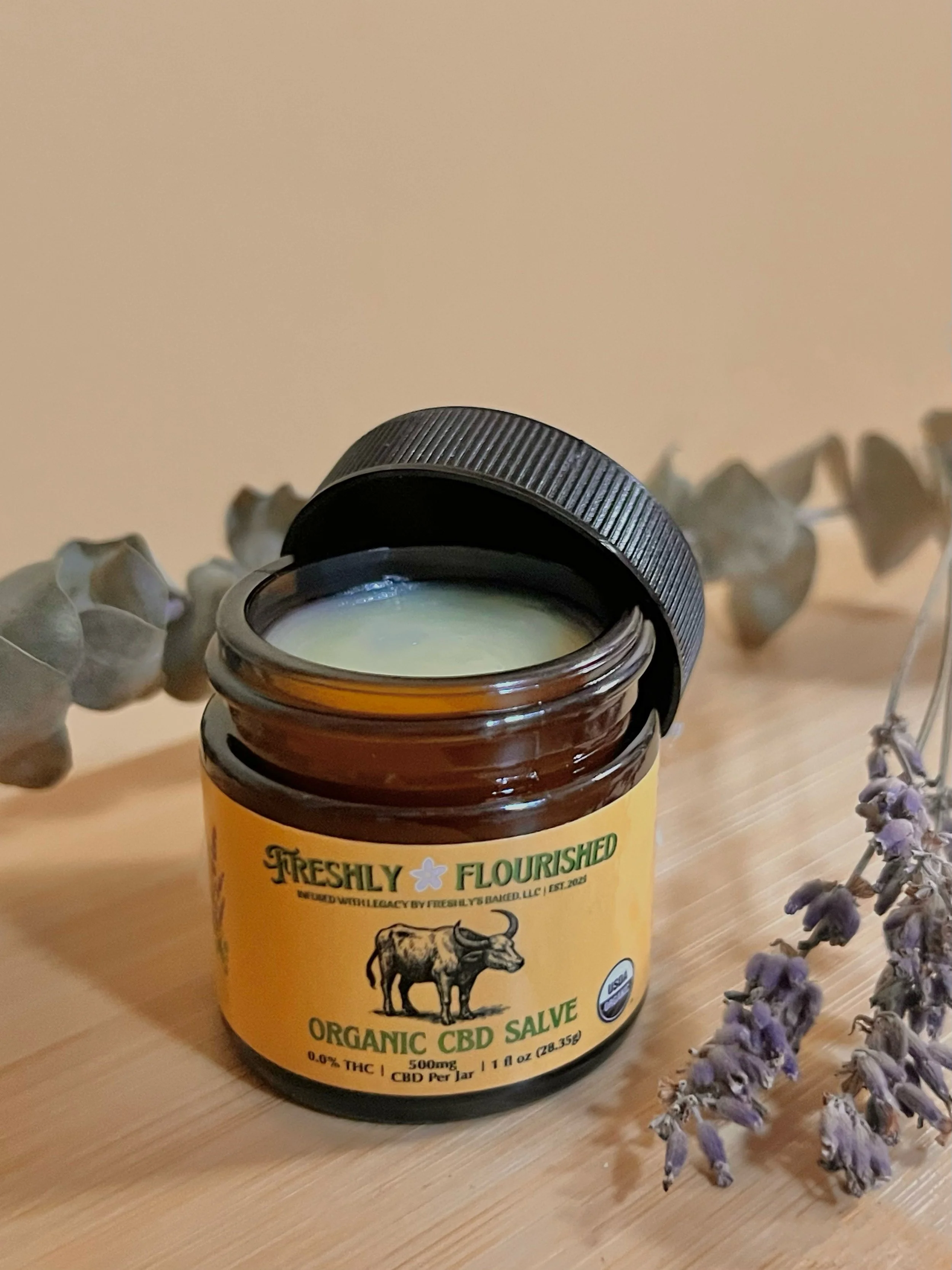 An open jar of organic CBD salve labeled "Freshly Flourished" with lavender flowers and eucalyptus beside it on a wooden surface.