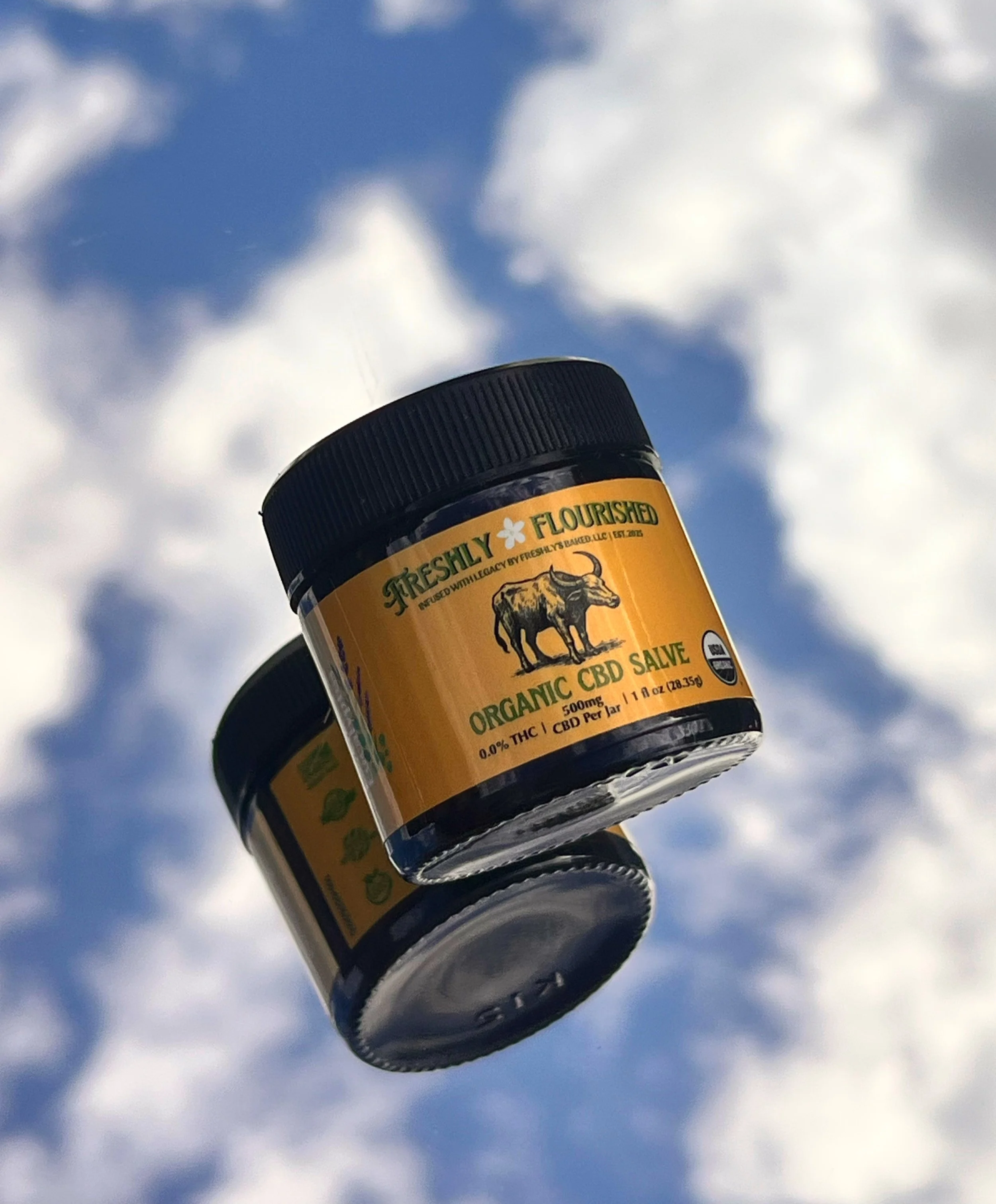 A small container of organic CBD salve with a yellow label featuring a carabao, of water buffalo, illustration, held up against a cloudy blue sky.