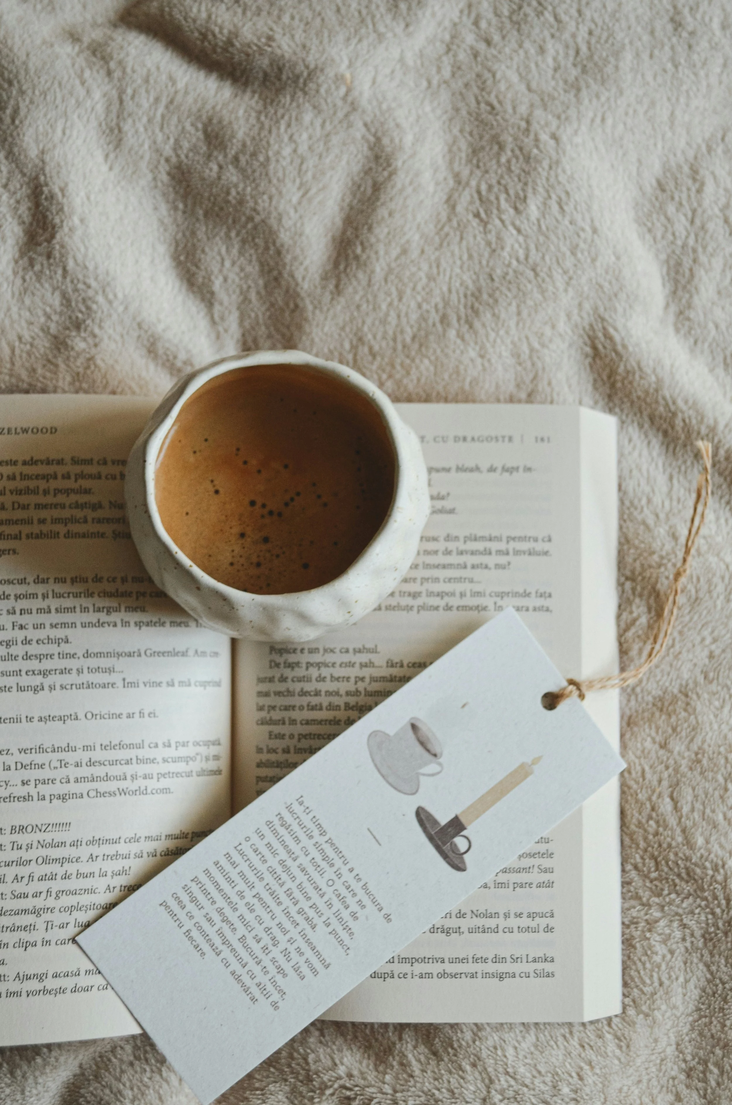 A cup of coffee or espresso placed on an open book and a white paper with a small illustration of a microscope, a magnifying glass, and a pencil, all on a soft beige textured fabric.