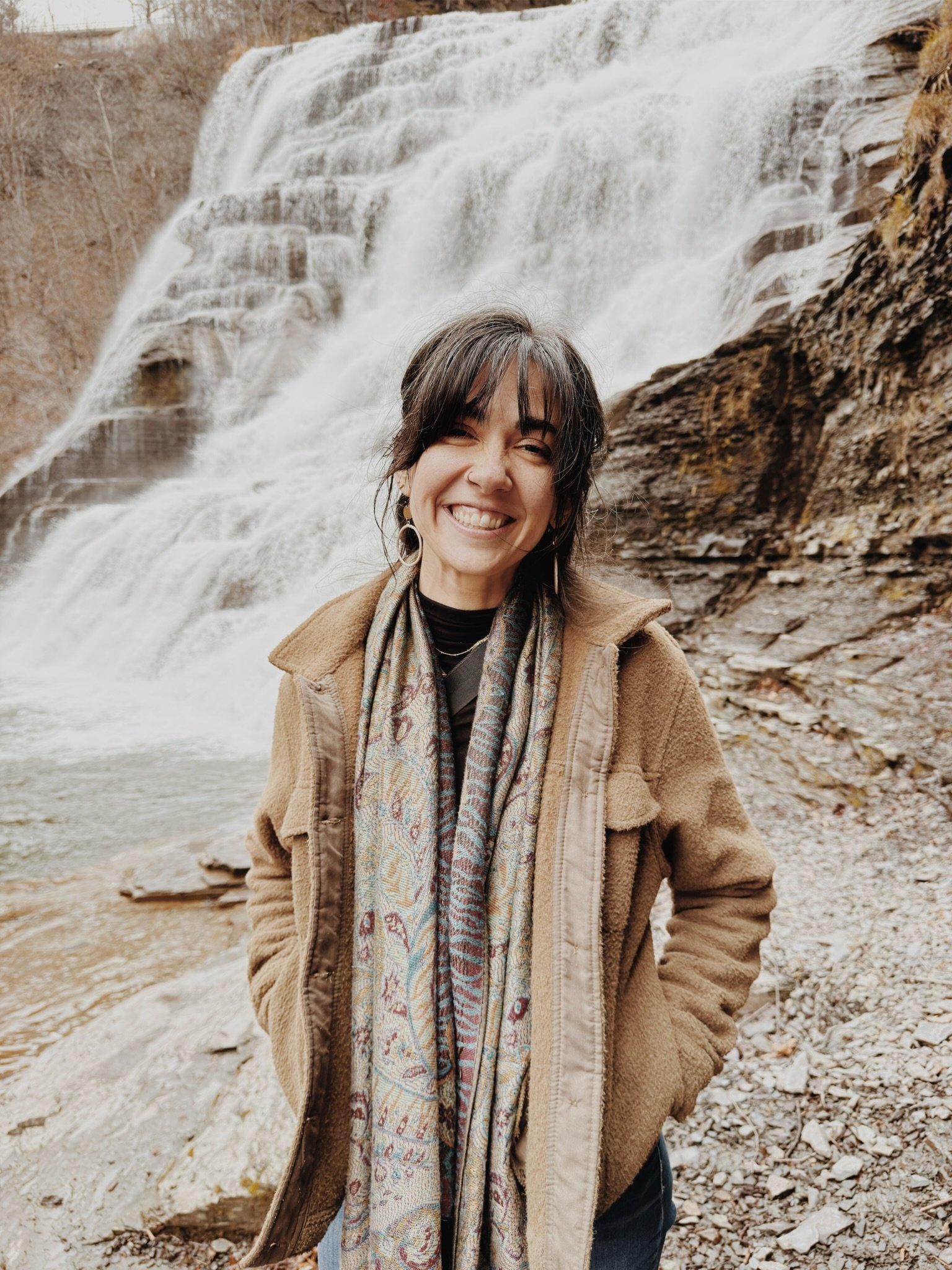 A young woman with dark hair, wearing a tan hoodie and a patterned scarf, smiling in front of a large waterfall and rocky landscape.