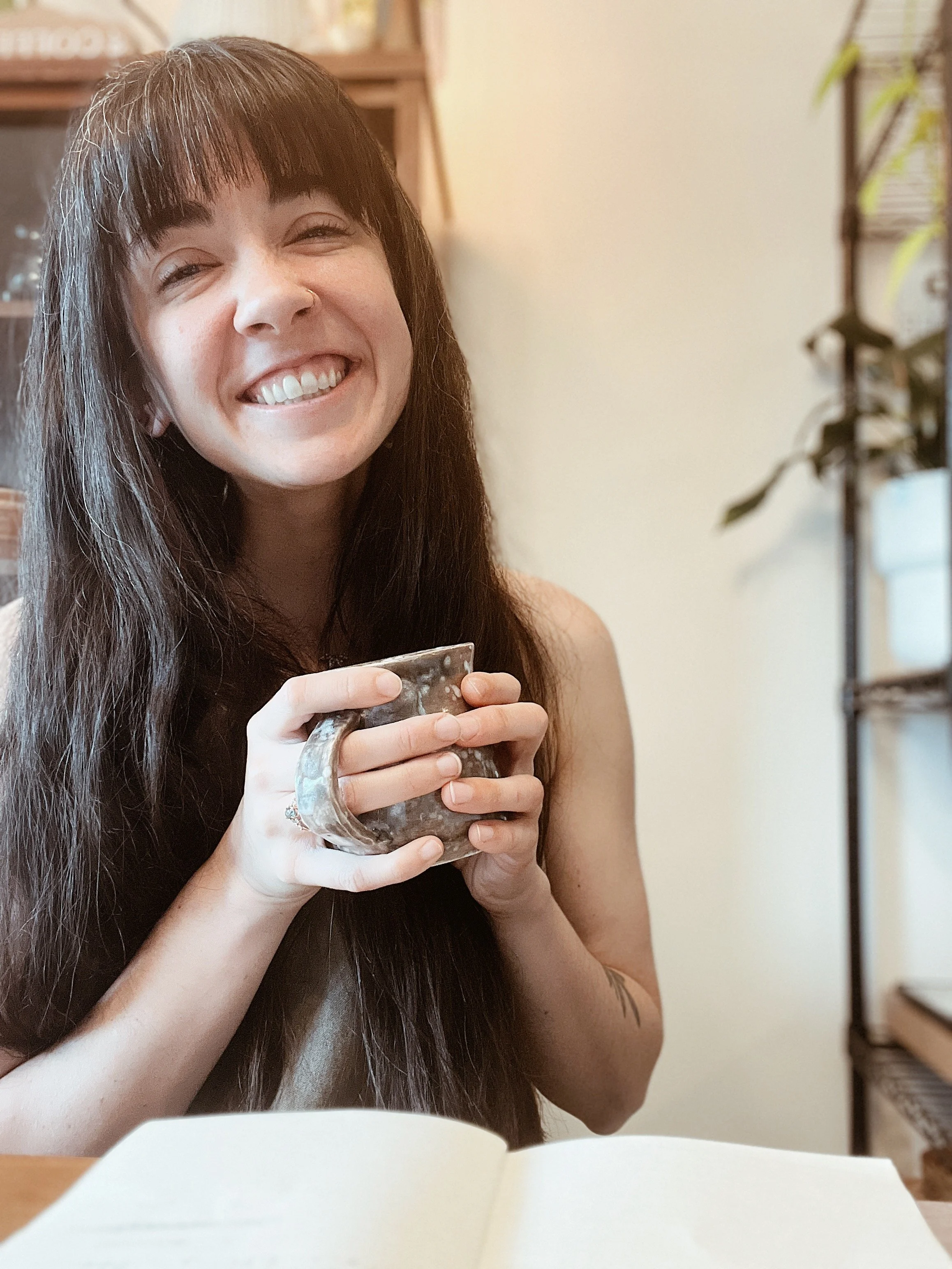 A woman with long brown hair, a nose ring, and a tattoo on her arm, smiling and holding a cup, sitting at a table with an open notebook in front of her in a cozy indoor setting.