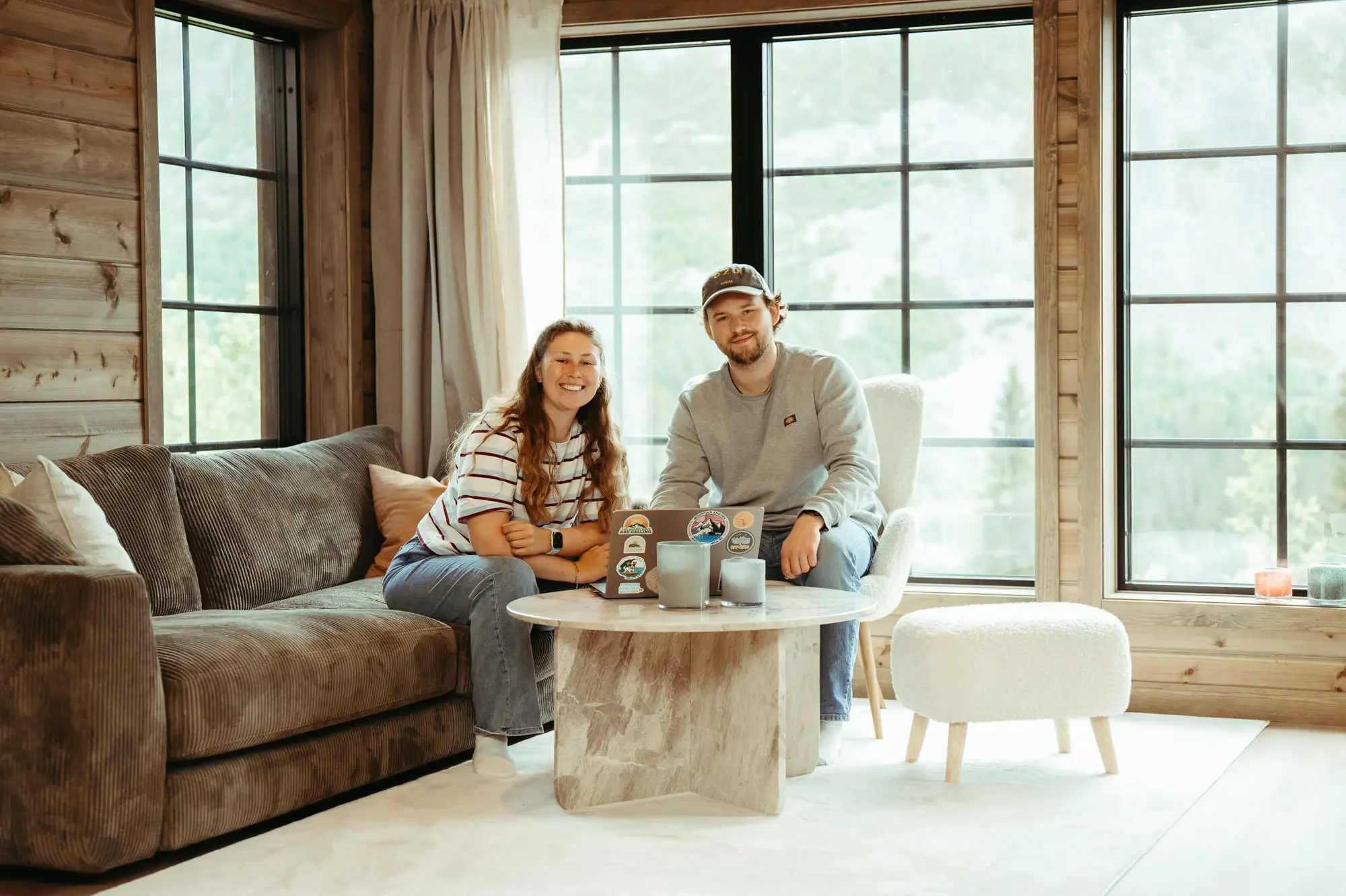 Två personer i ett mysigt vardagsrum, sittandes på en soffa med ett runt soffbord framför dem, med stora fönster i bakgrunden och mycket naturligt ljus, och dekorativa ljus på bordet.