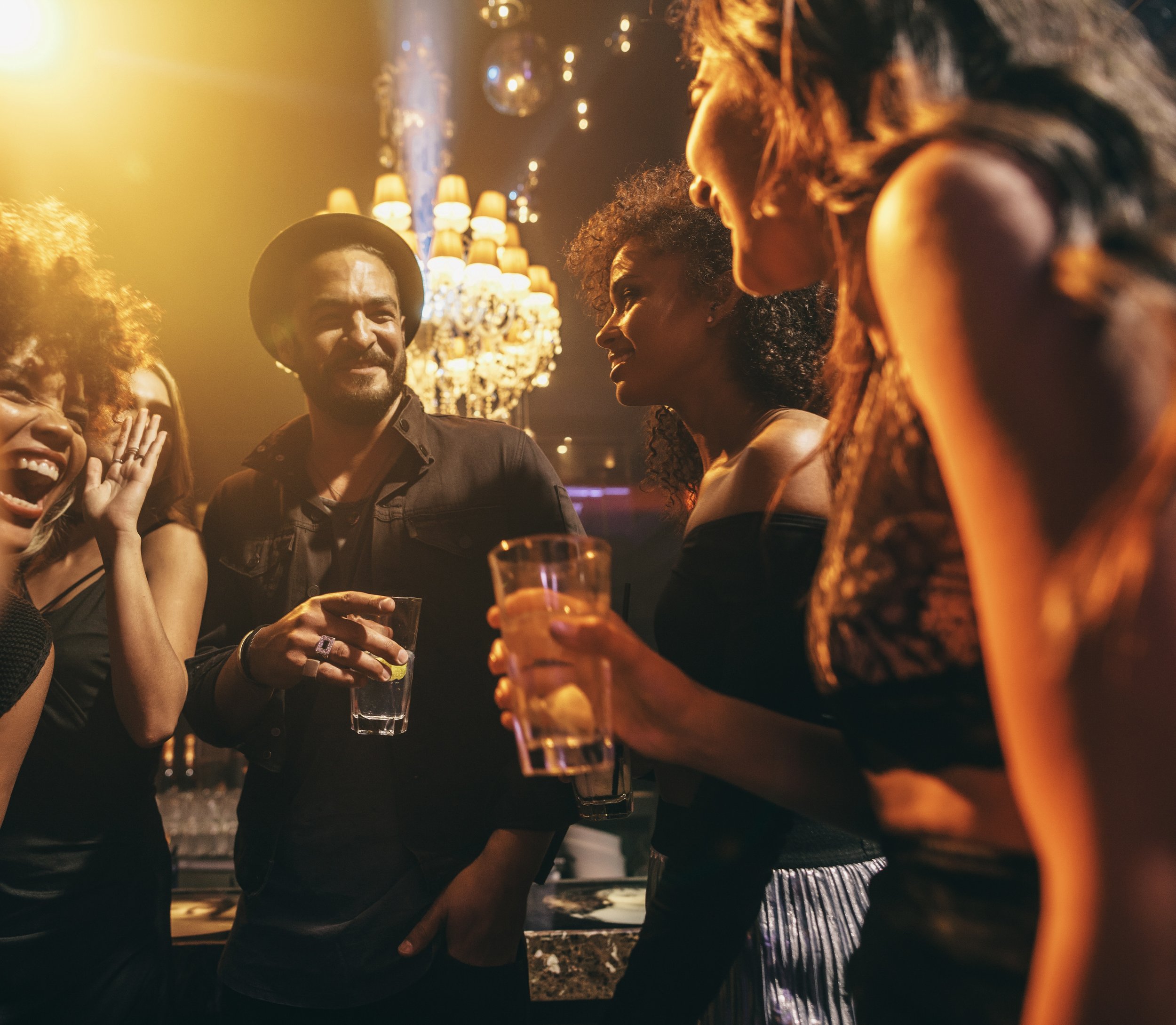 A group of friends enjoying a night out at a bar or club, holding drinks, and engaging in conversation with warm lighting and a chandelier in the background.