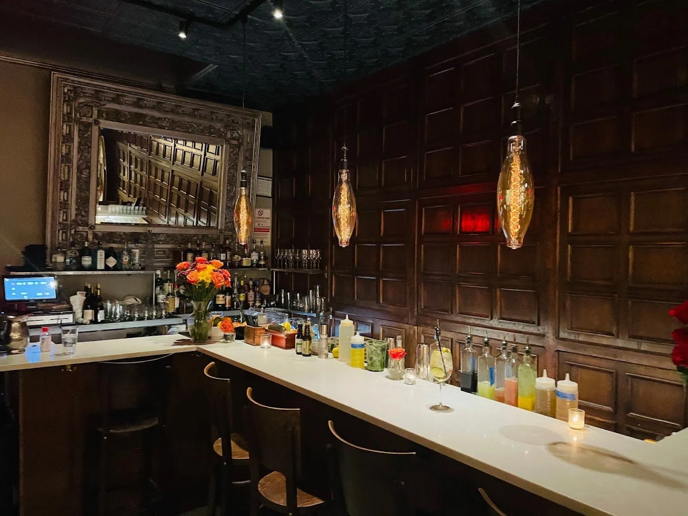 A dimly lit bar area with a white counter, decorated with flowers, bottles, and glasses. Three hanging pendant lights with gold accents illuminate the space. There is a large ornate mirror with a reflective surface on the wall behind the bar, showing a slanted view of a staircase. The dark wood-paneled wall adds to the elegant, cozy ambiance.
