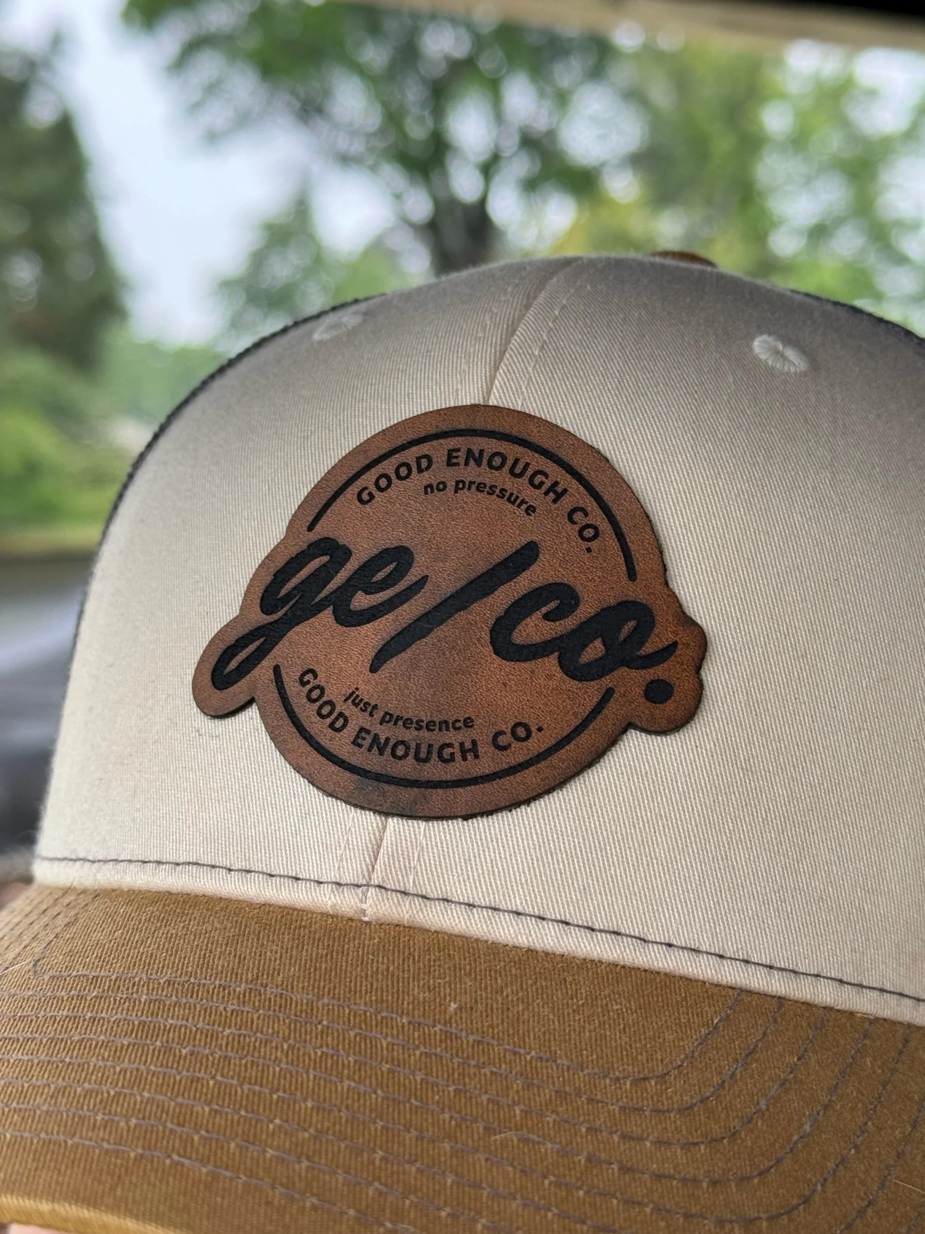 A beige baseball cap with a brown leather patch on the front that reads, "Good Enough Co. Just Presence, Good Enough," against a background of blurred green and white trees.