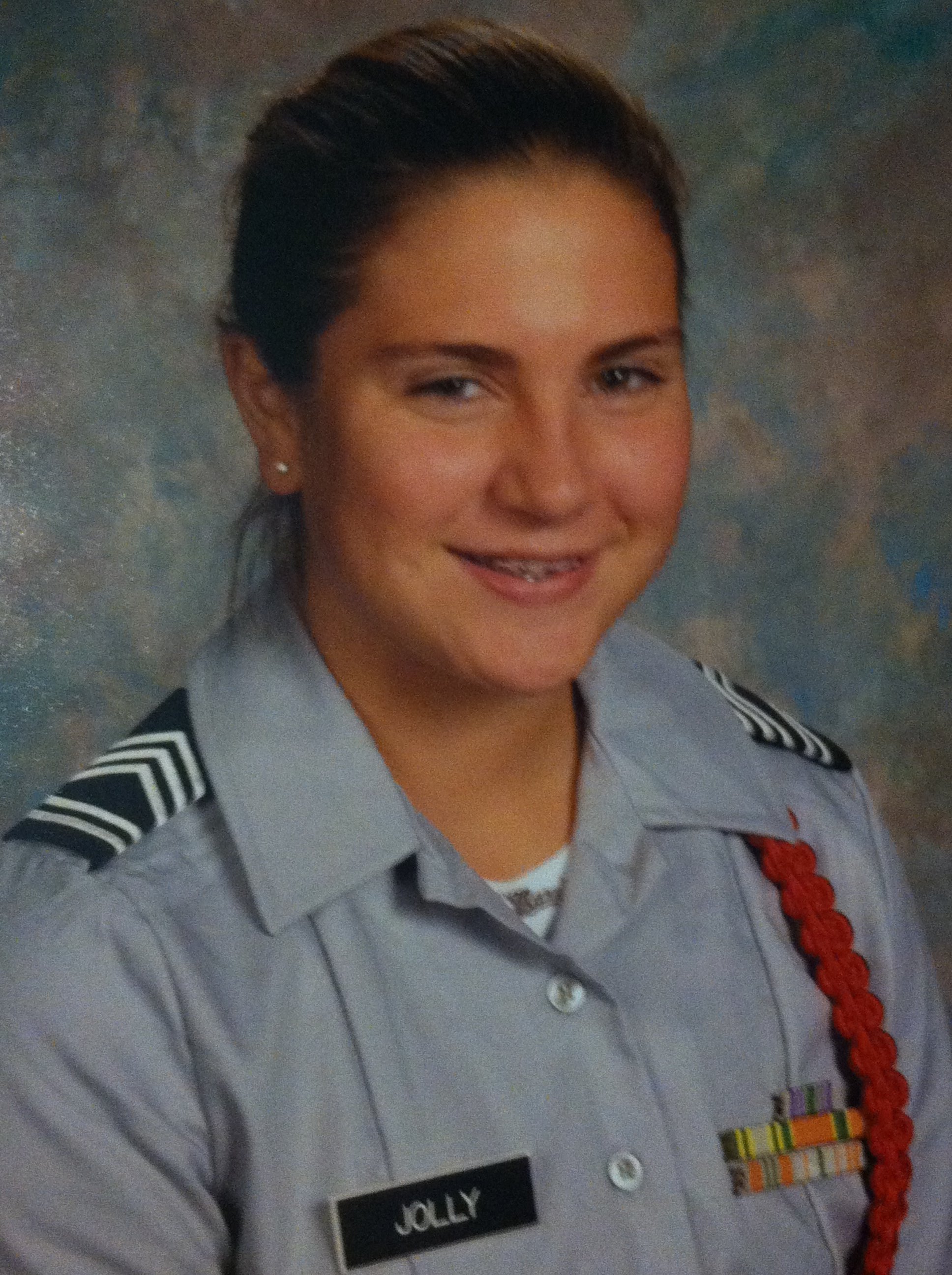 Close-up of a woman in a military uniform with a name tag that says 'JOLLY.' She has dark hair, is wearing pearl earrings, and is smiling.