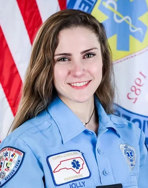 Young woman in EMT uniform smiling, standing in front of American flag and emergency services flag.