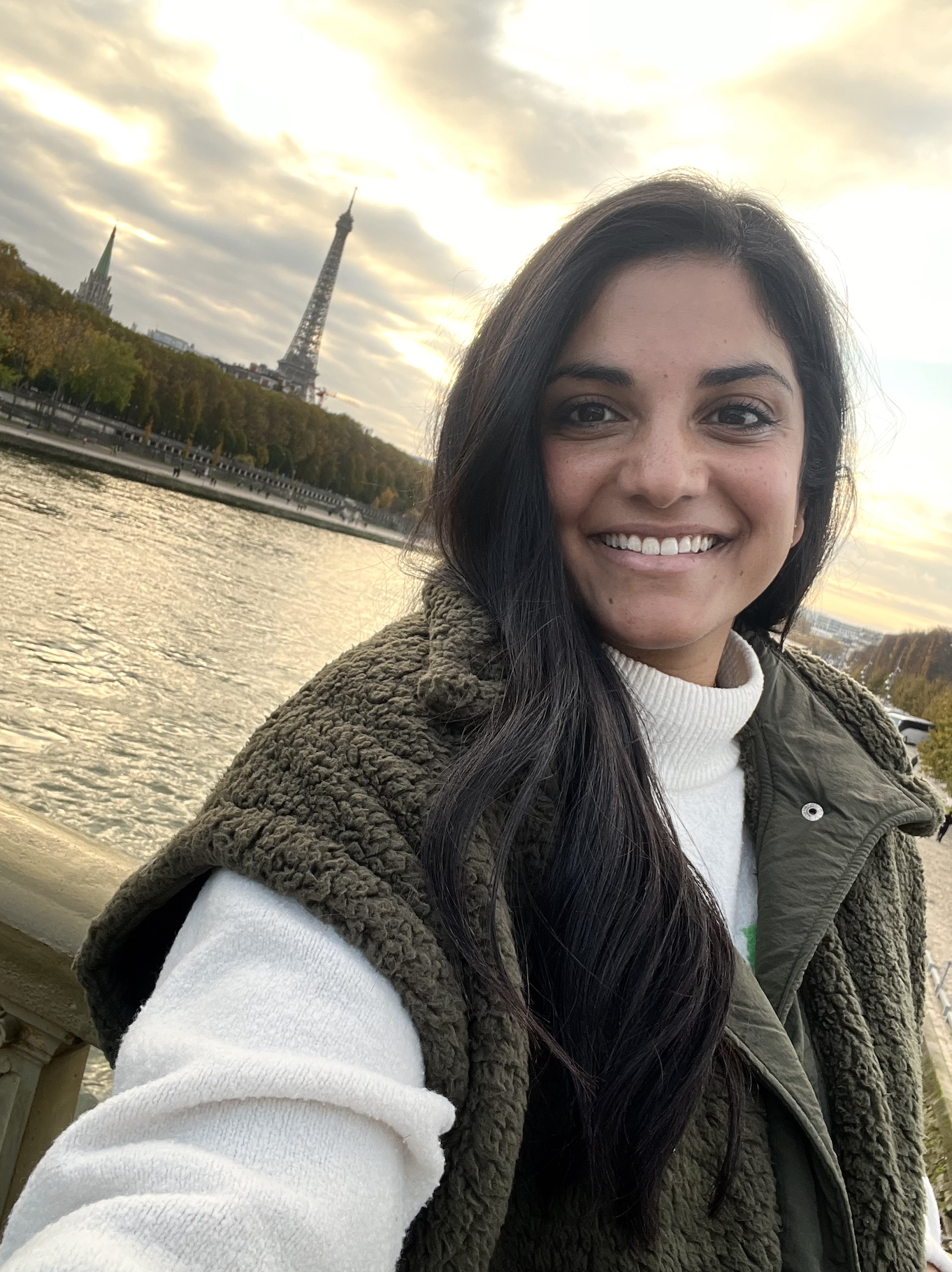 A woman with long dark hair smiling for a selfie near a river with the Eiffel Tower in the background at sunset.