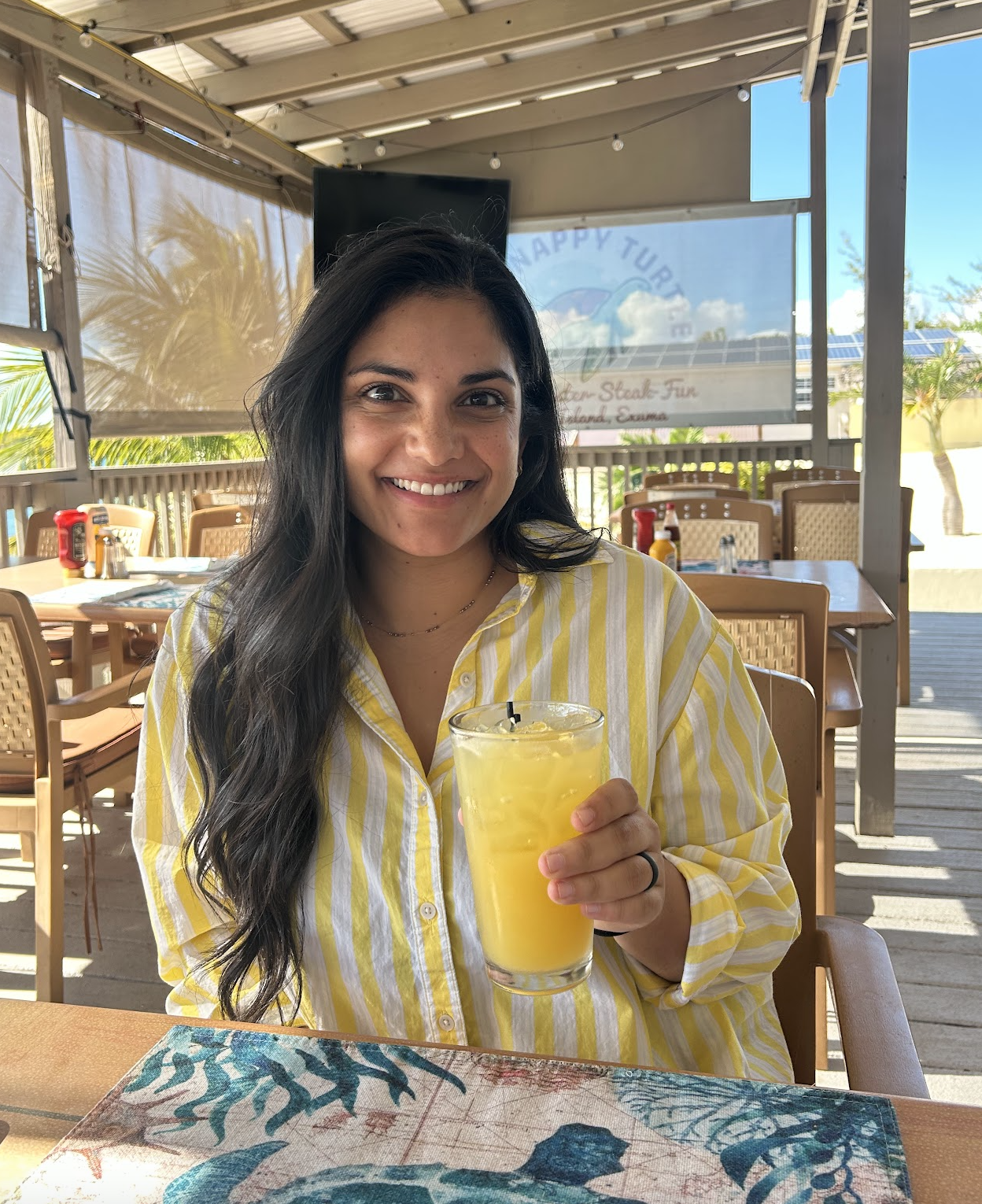 A woman in a yellow and white striped shirt sitting at an outdoor restaurant, holding a yellow drink with a straw, smiling at the camera. The restaurant has wooden tables and chairs, with palm trees and blue sky visible in the background.