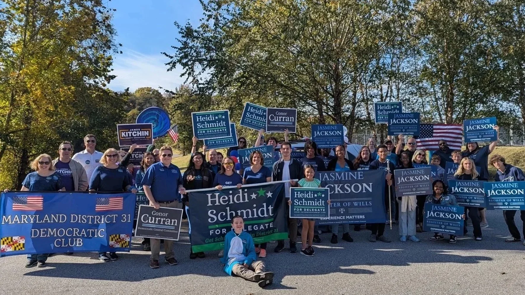 Group of people holding campaign signs for Heidi Schmidt, Jasmine Jackson, and Conor Curran, standing outdoors with trees and a blue sky in the background.