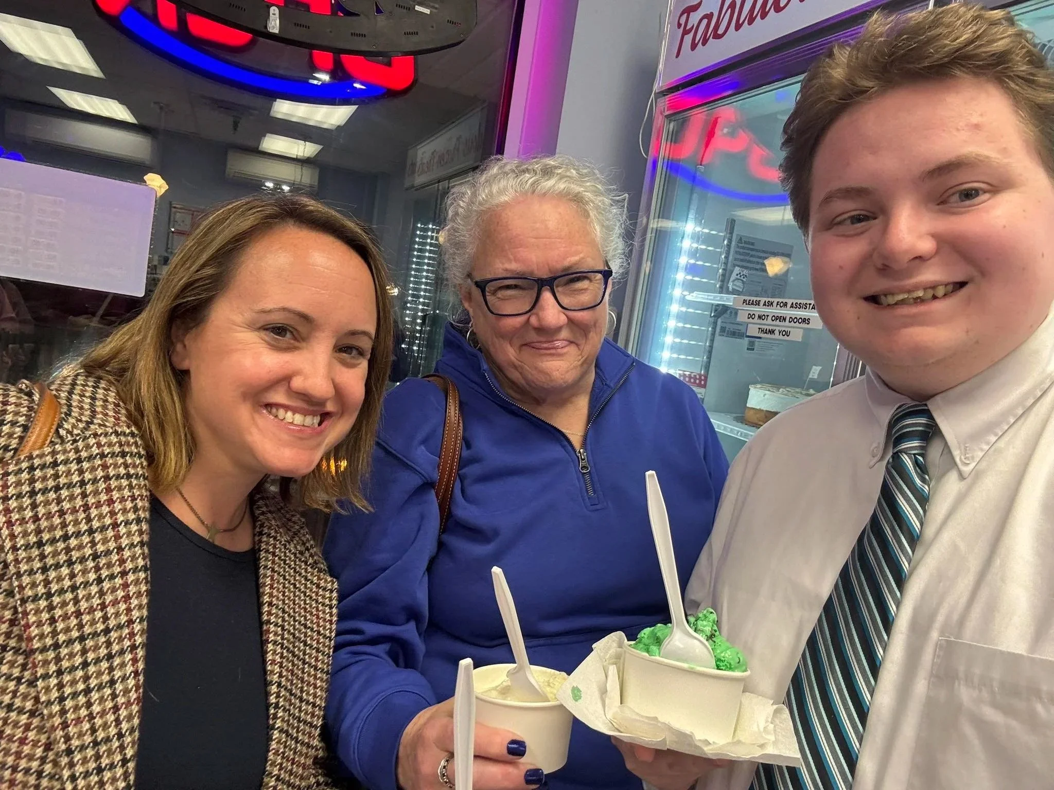 Three people sharing a treat of ice cream at a Dairy Queen store with a neon sign in the background. Two women and a young man, all smiling, are holding cups of ice cream with spoons.