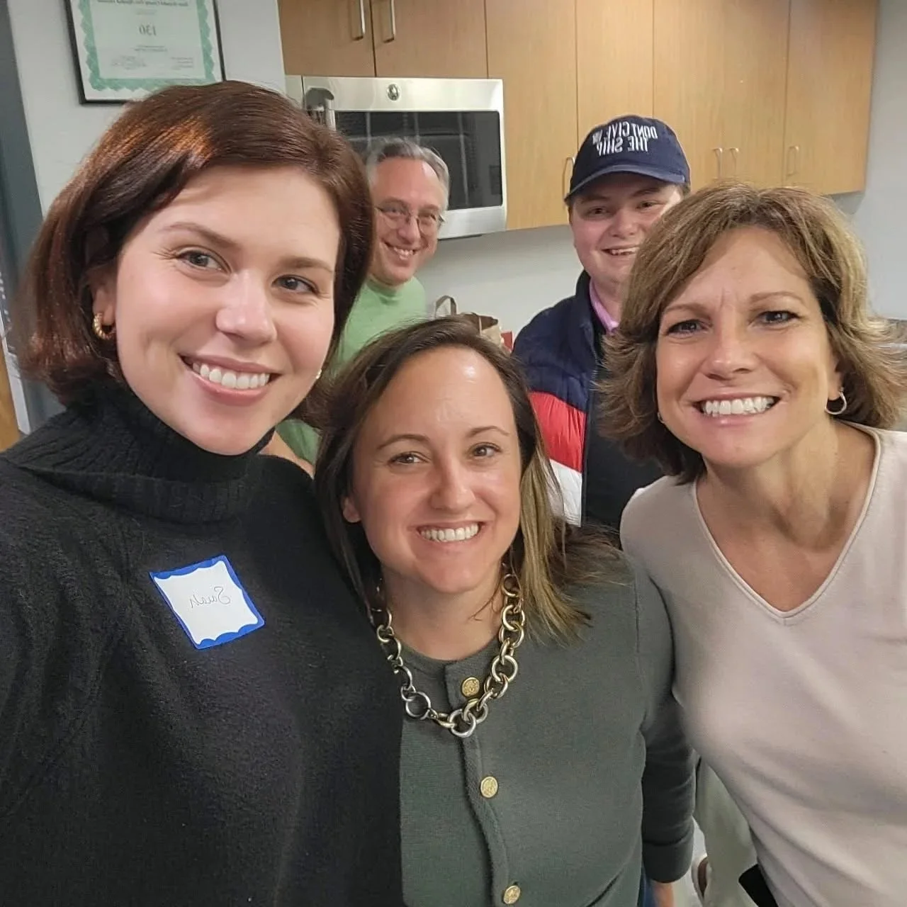 A group of five adults smiling and taking a selfie in an indoor setting, with kitchen cabinets and a microwave in the background.