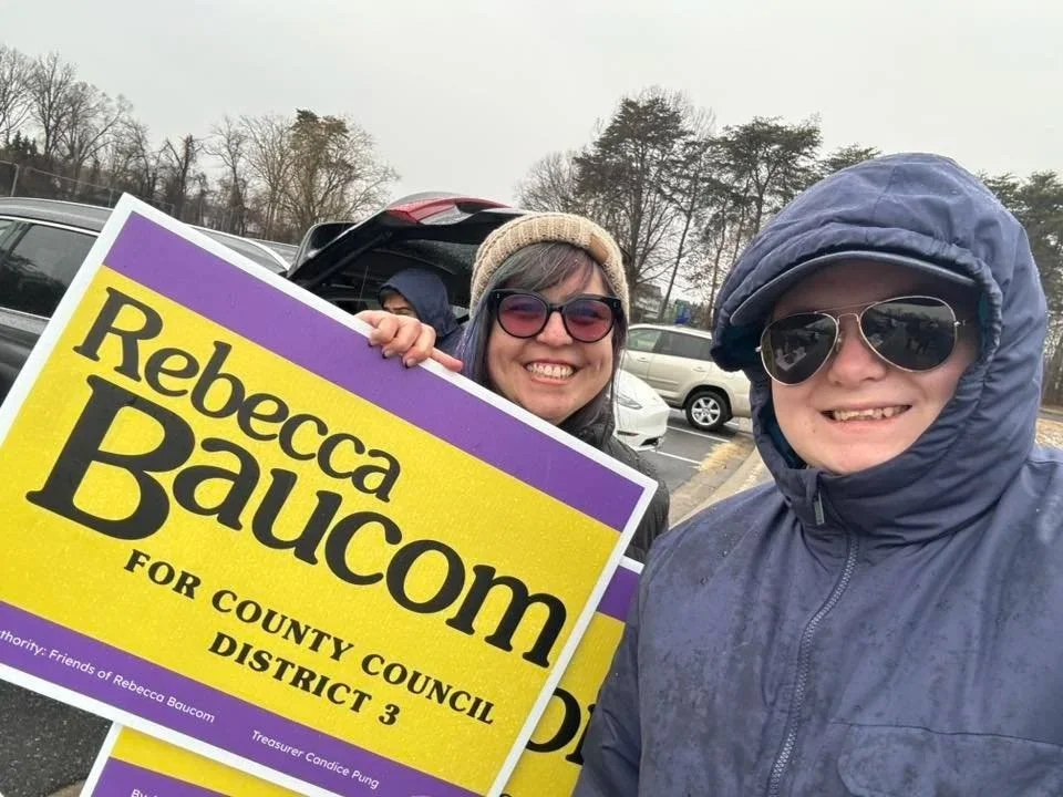 Two women outdoors, one holding an election campaign sign for Rebecca Baucom running for county council district 3, both wearing jackets and sunglasses, overcast weather with bare trees in the background.
