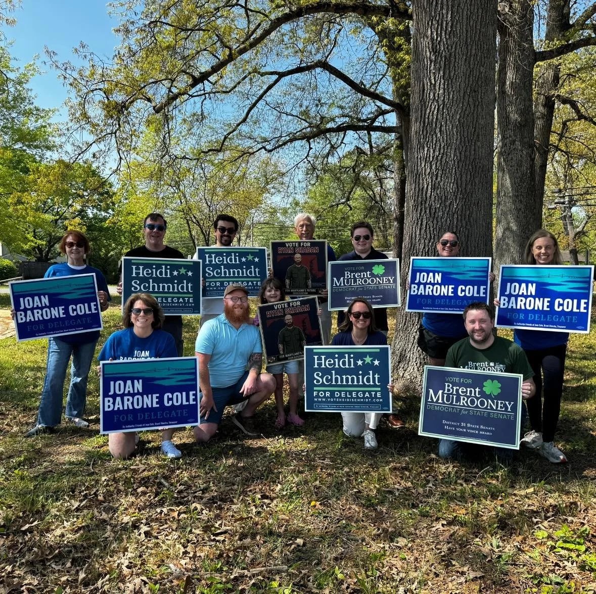 Group of people outdoors holding campaign signs for Heidi Schmidt, Joan Barone Cole, and Brent Mulrooney, standing near large trees on a sunny day.