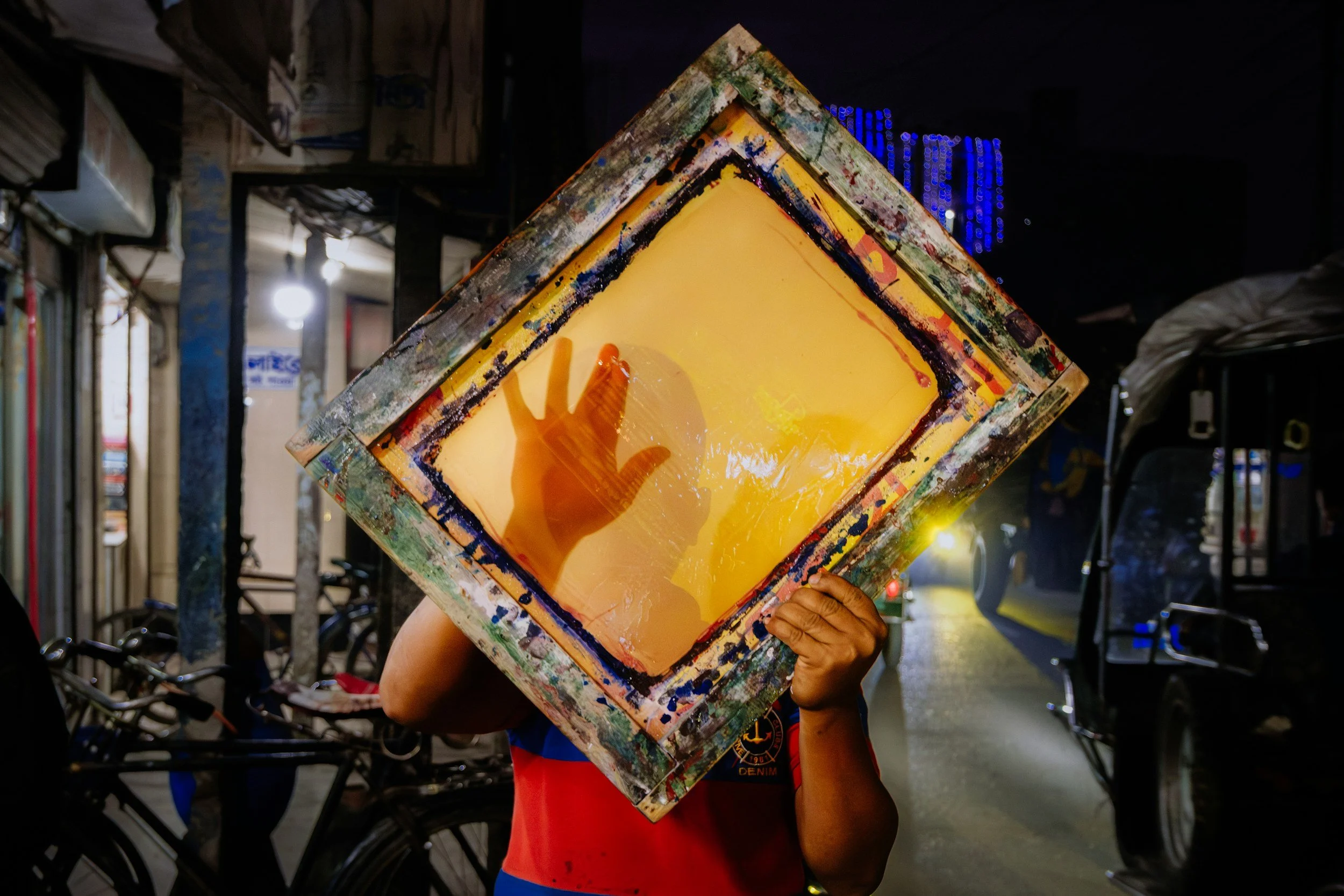 Person holding a colorful abstract painting in front of their face on a city street at night.