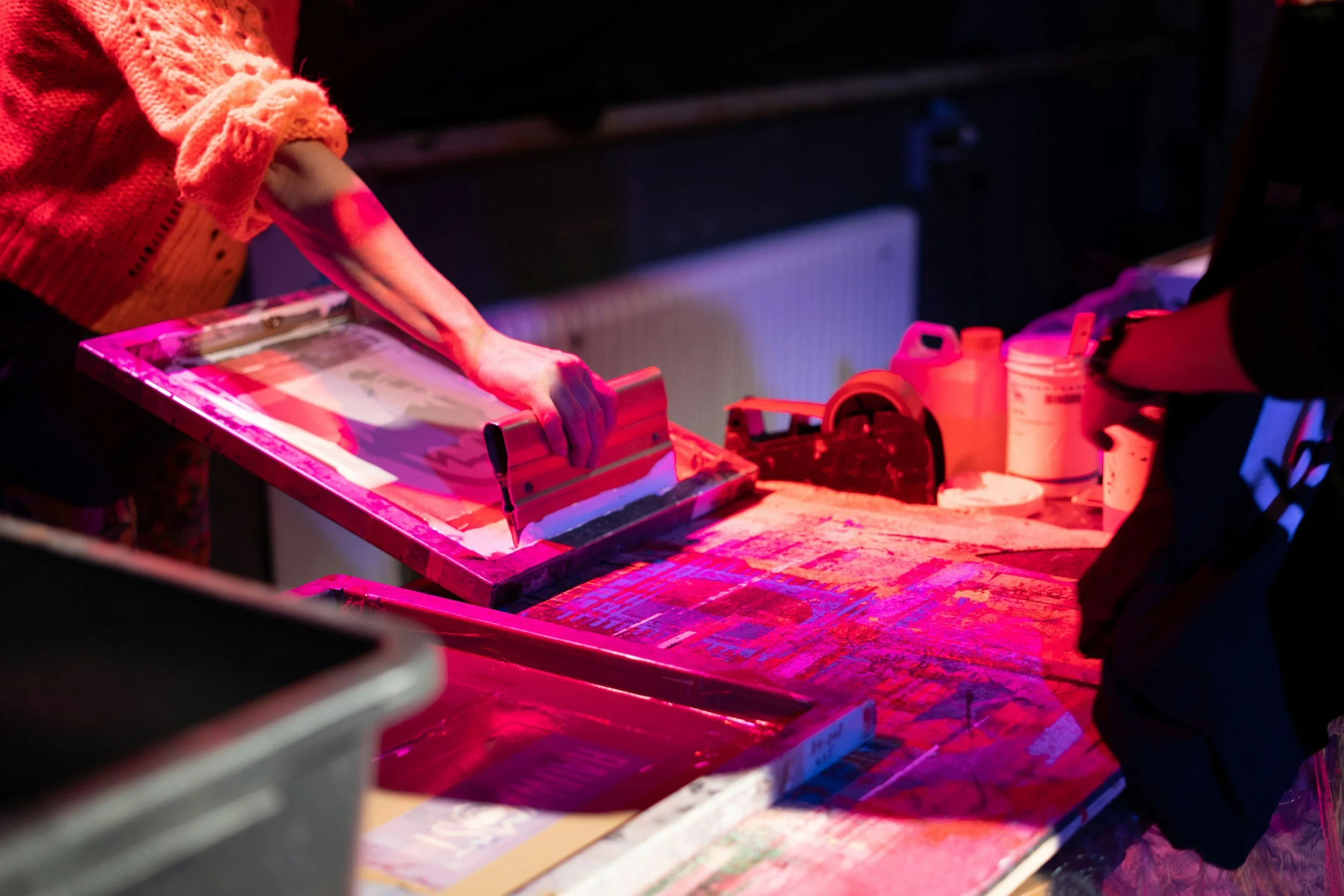 A person in an orange sweater working on a printing press or screen printing table in a dimly lit room with pink and purple lighting.