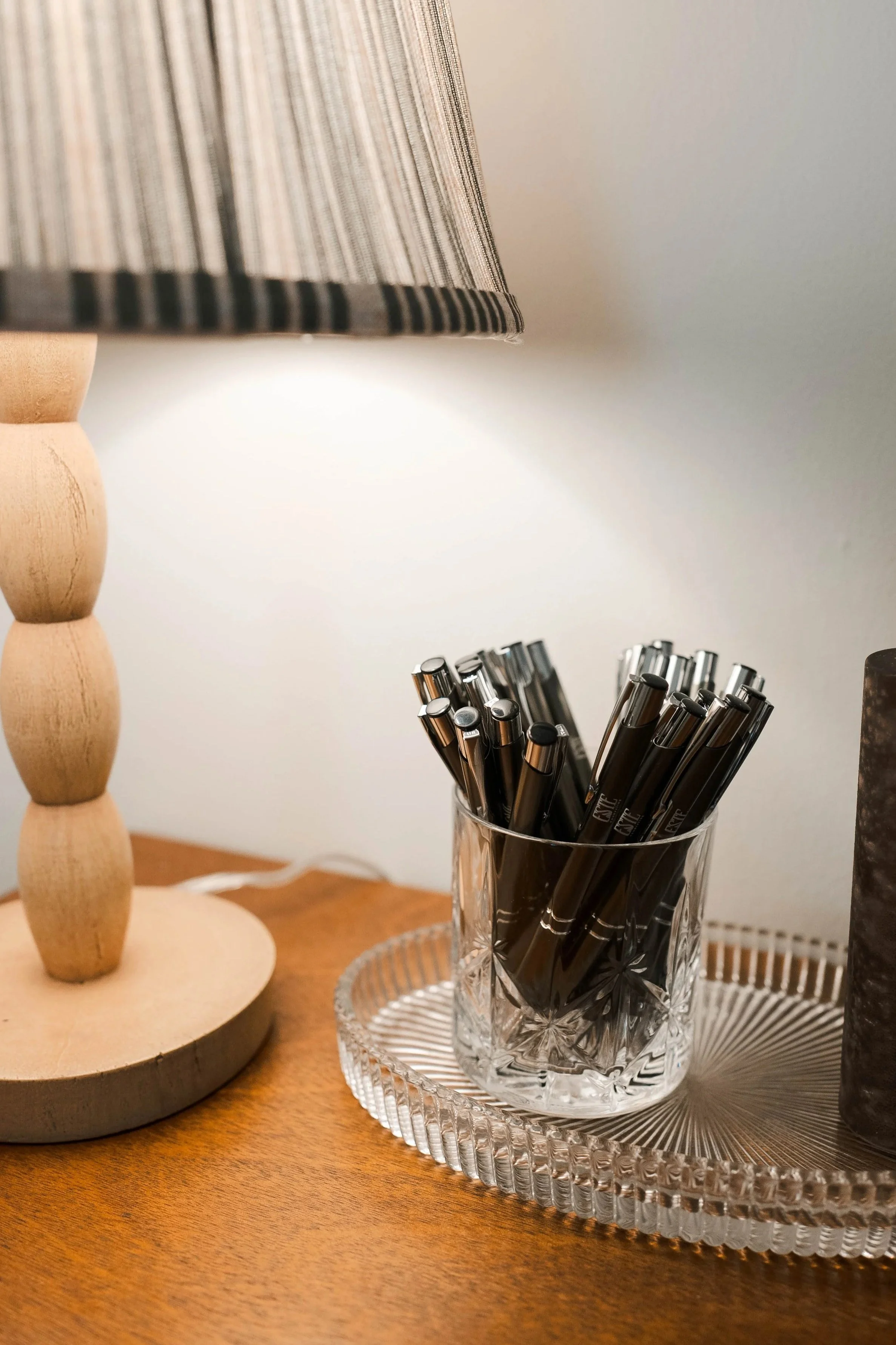 Black pens in a clear, textured glass holder on a wooden table, with a decorative glass tray underneath, a wooden lamp with a fabric shade to the left, against a plain wall.