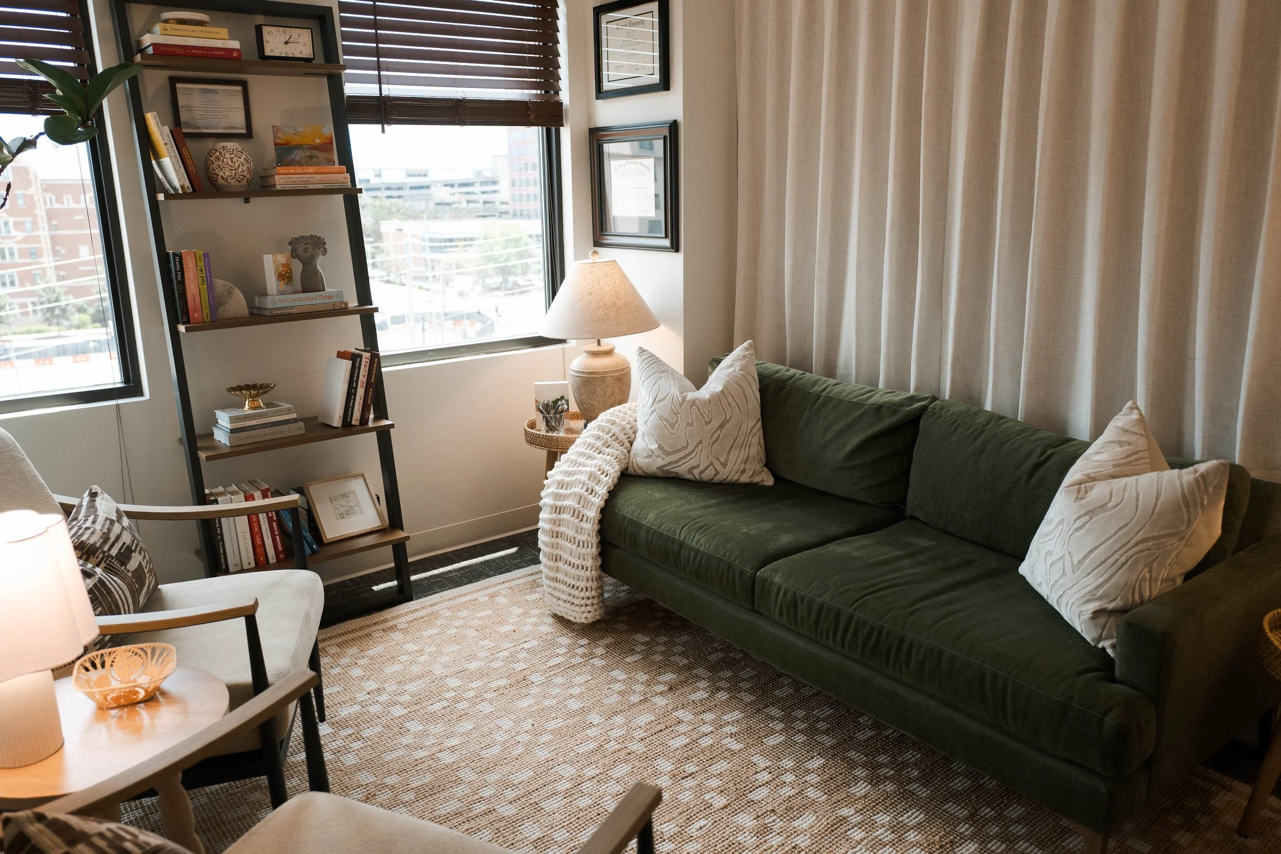 Living room corner with a green velvet sofa, pillows, and a white blanket. A side table with a lamp, and a bookshelf with books and decorative items. Large windows with wooden blinds letting in natural light, framed certificates or artwork on the wall, and a patterned beige carpet.