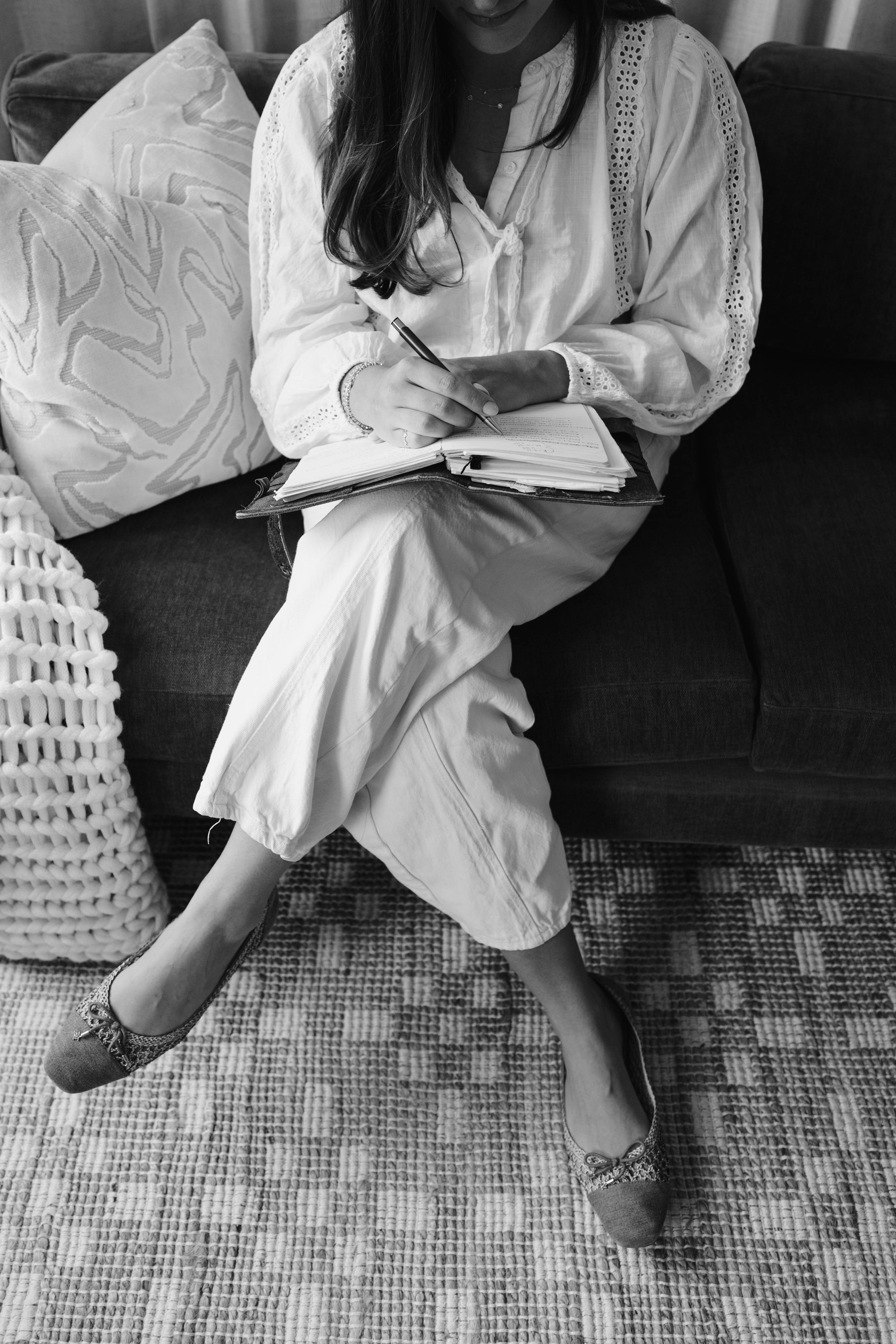 A woman sitting on a couch, writing in a notebook with a pen. She is wearing a white blouse with embroidery details and light-colored pants. Her legs are crossed, and she is wearing a ring, bracelet, and patterned shoes. There are decorative pillows behind her and a rug beneath her on the floor.