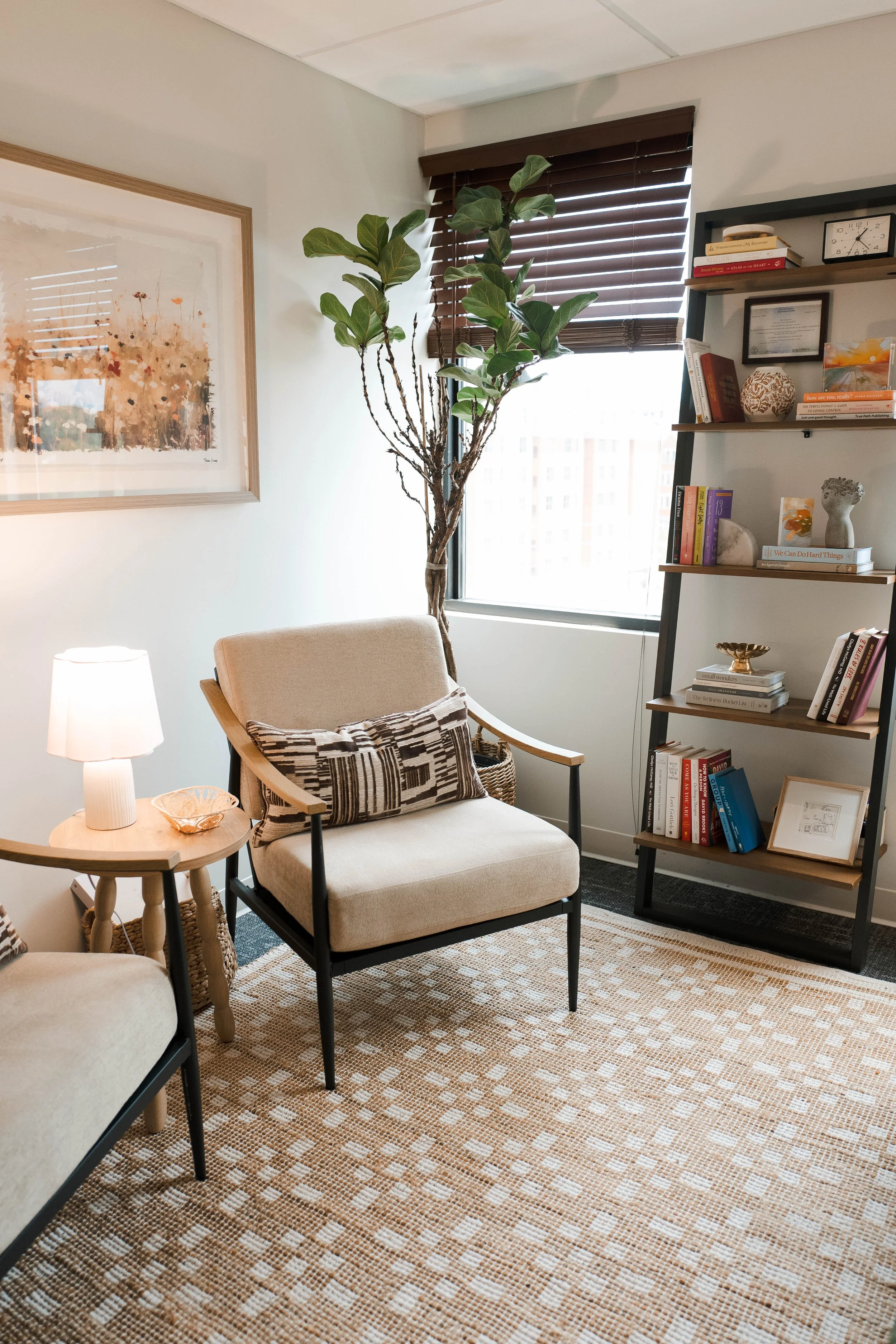 A cozy living room corner with a beige armchair, black metal legs, and a patterned cushion. Next to it is a small round wooden side table with a white lamp and a decorative bowl. There is a large plant with multiple branches and broad green leaves near a window with wooden blinds. On the right, a black shelf displays books, framed pictures, a clock, and decorative items. A colorful abstract painting hangs on the wall to the left. The floor has a patterned beige and brown area rug.