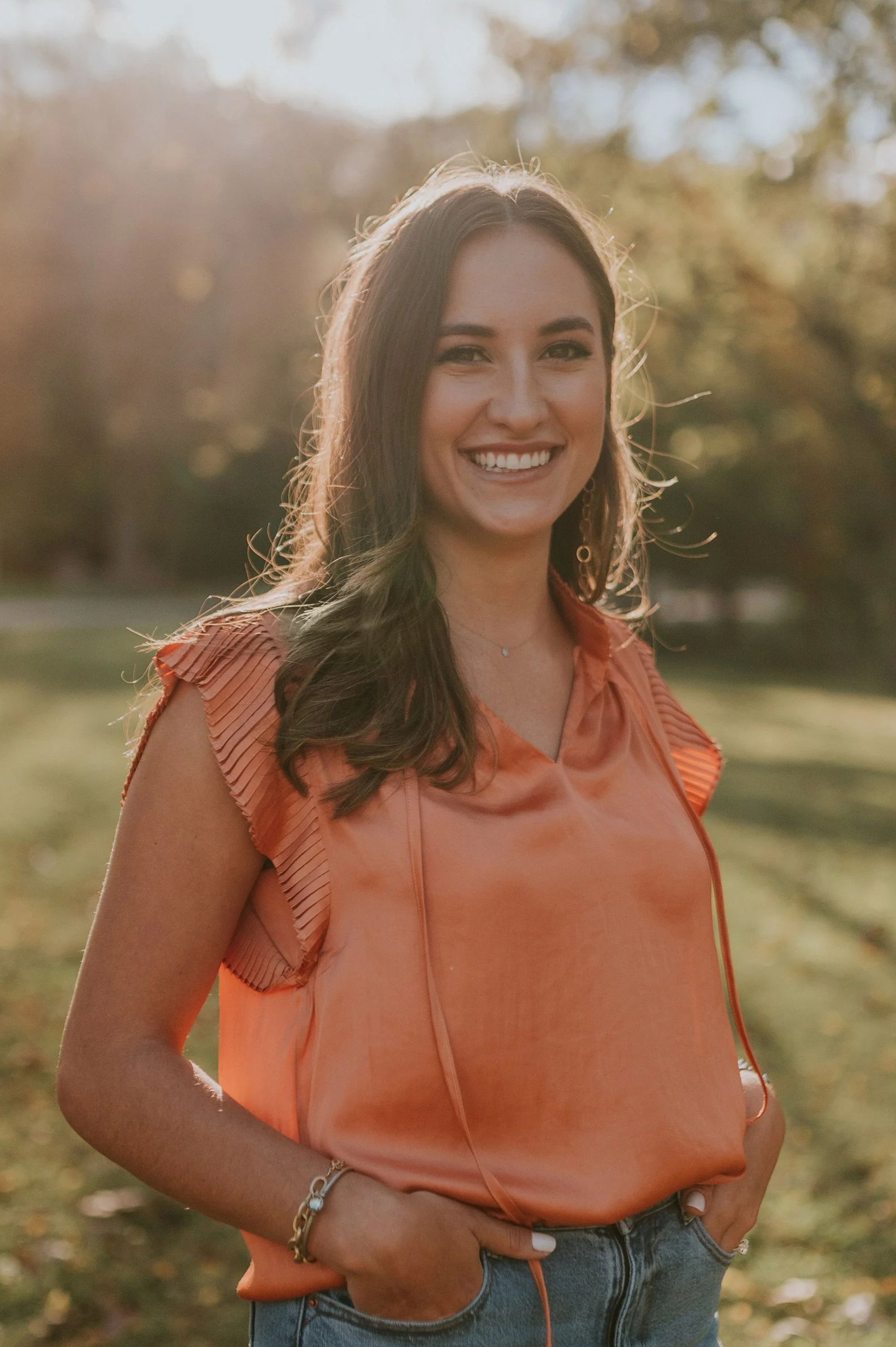 A young woman smiling outdoors during sunset, wearing an orange top and jeans, with trees and blurred nature in the background.