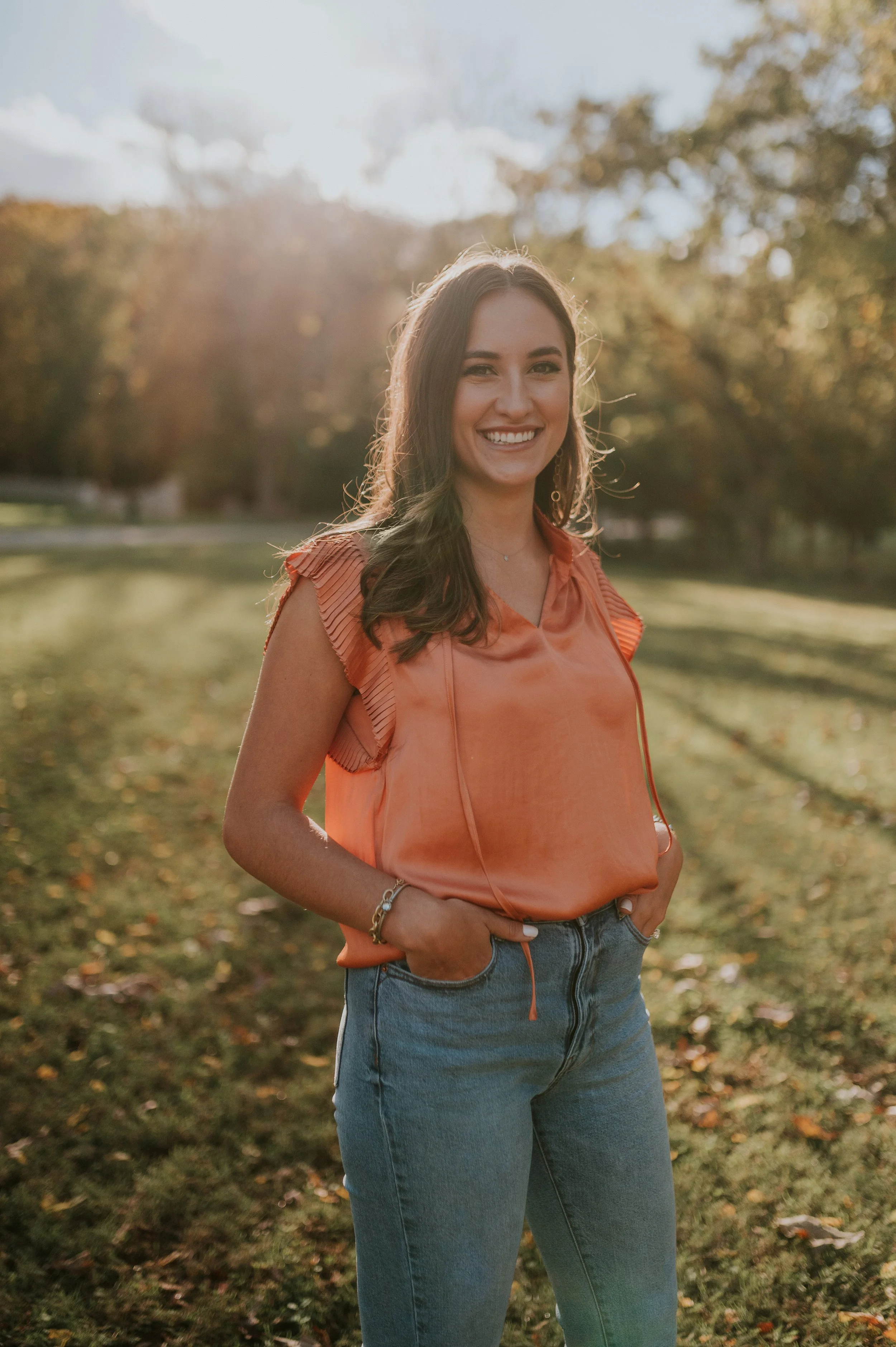 A young woman smiling outdoors in a park during fall, wearing an orange top and blue jeans, with her hands in her pockets.