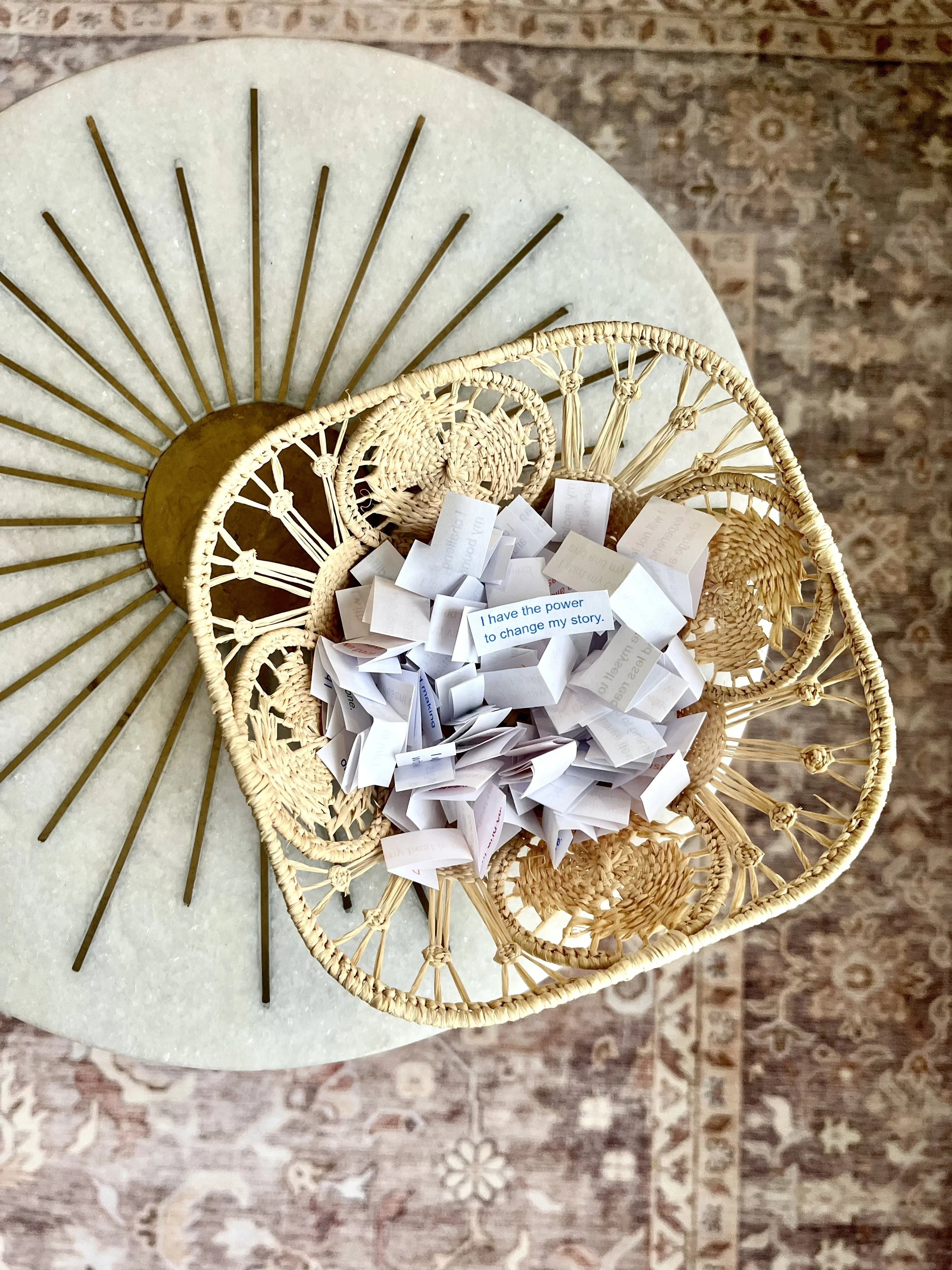 A wicker basket filled with small folded papers on a round table with a detailed patterned rug underneath.