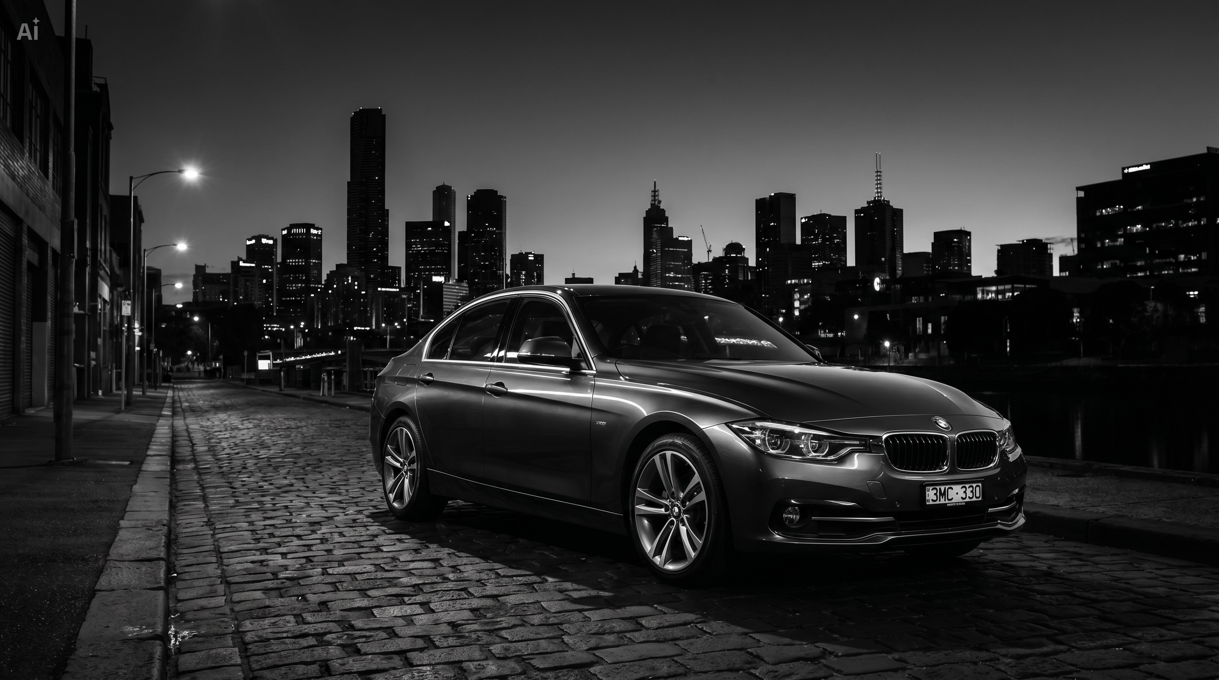 A black BMW sedan parked on a cobblestone street at night, with a city skyline of tall buildings in the background.