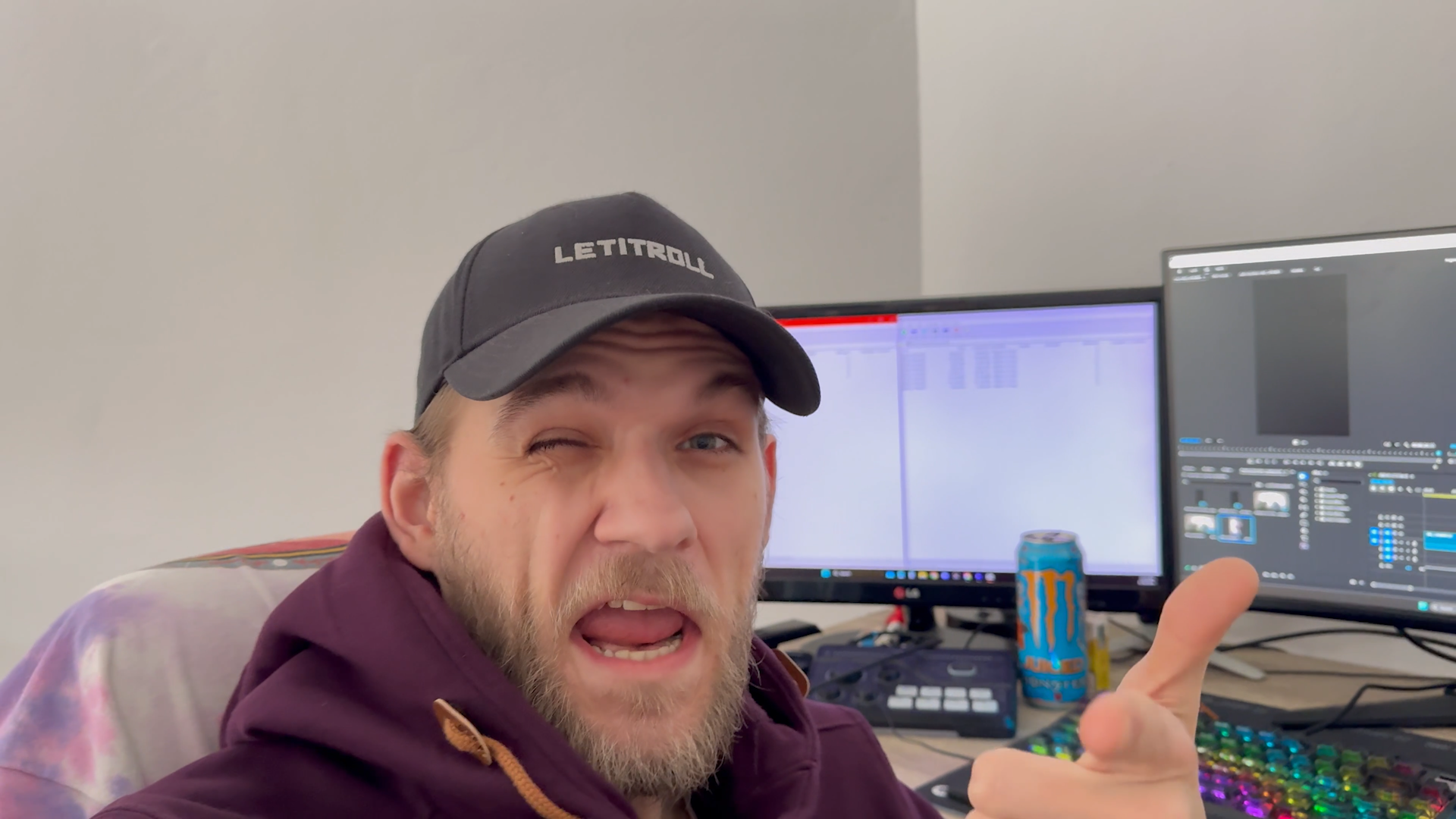 A man in a baseball cap and hoodie making a playful face, winking and sticking out his tongue, in front of dual computer monitors with video editing software visible.