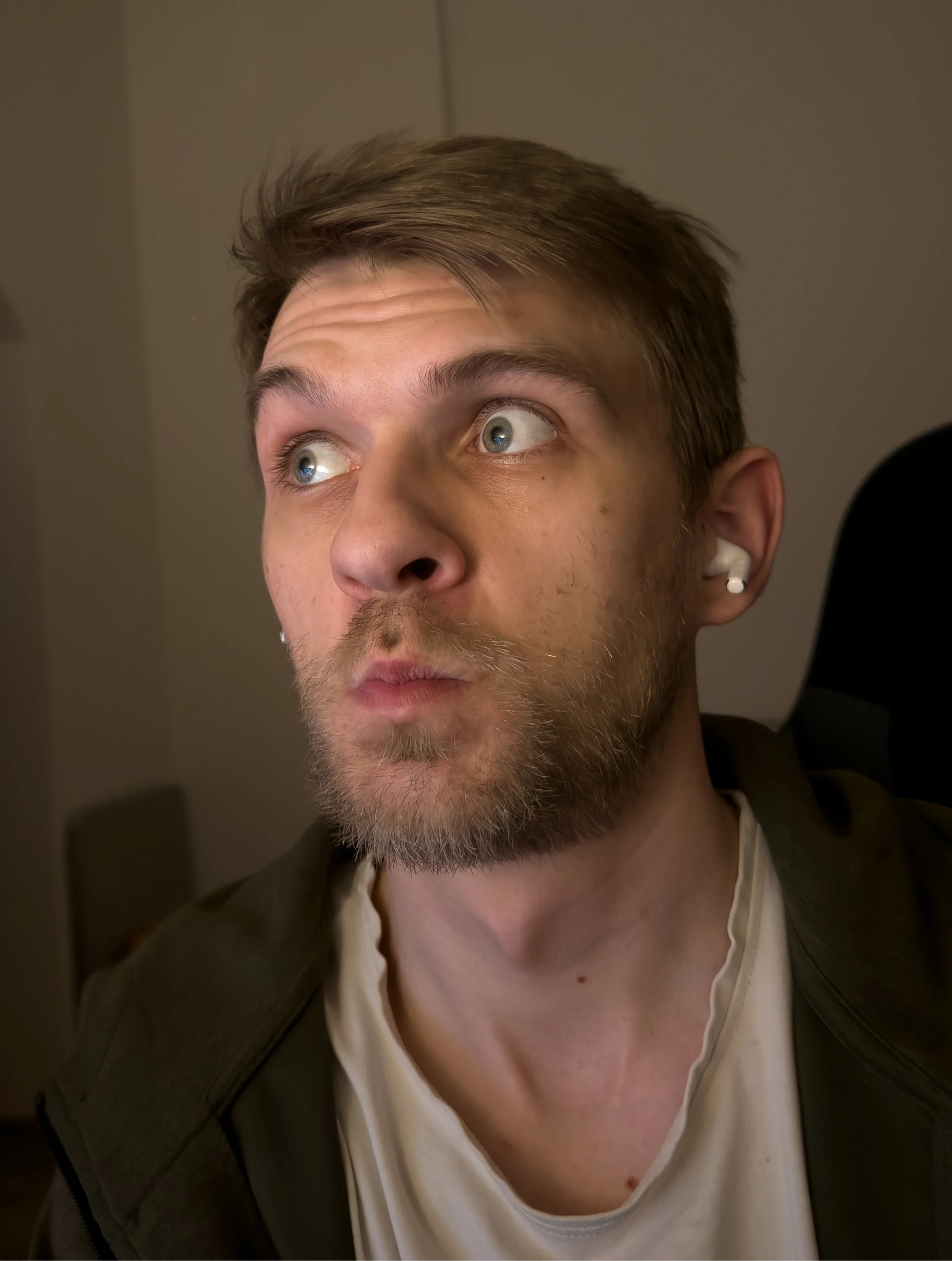 Close-up of a young man with light brown hair, blue eyes, and light facial hair, wearing white earbuds, looking to his right, in an indoor setting.